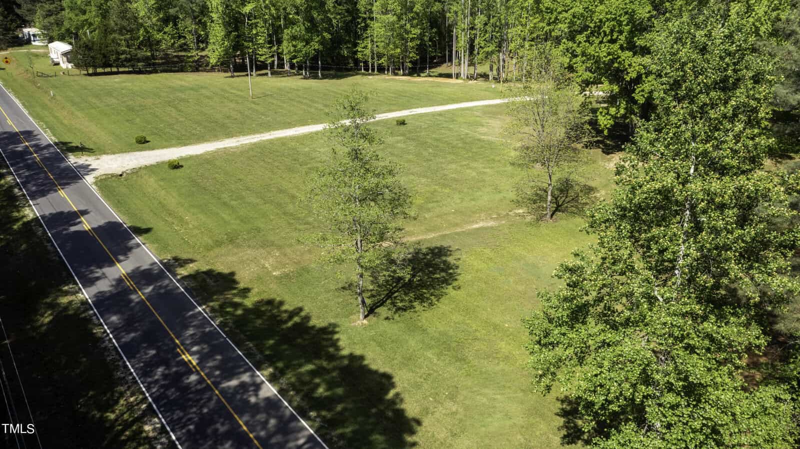 Aerial view of a grassy landscape with a long driveway leading to a white structure in the background. The scene includes a road on the left and clusters of trees surrounding the area.
