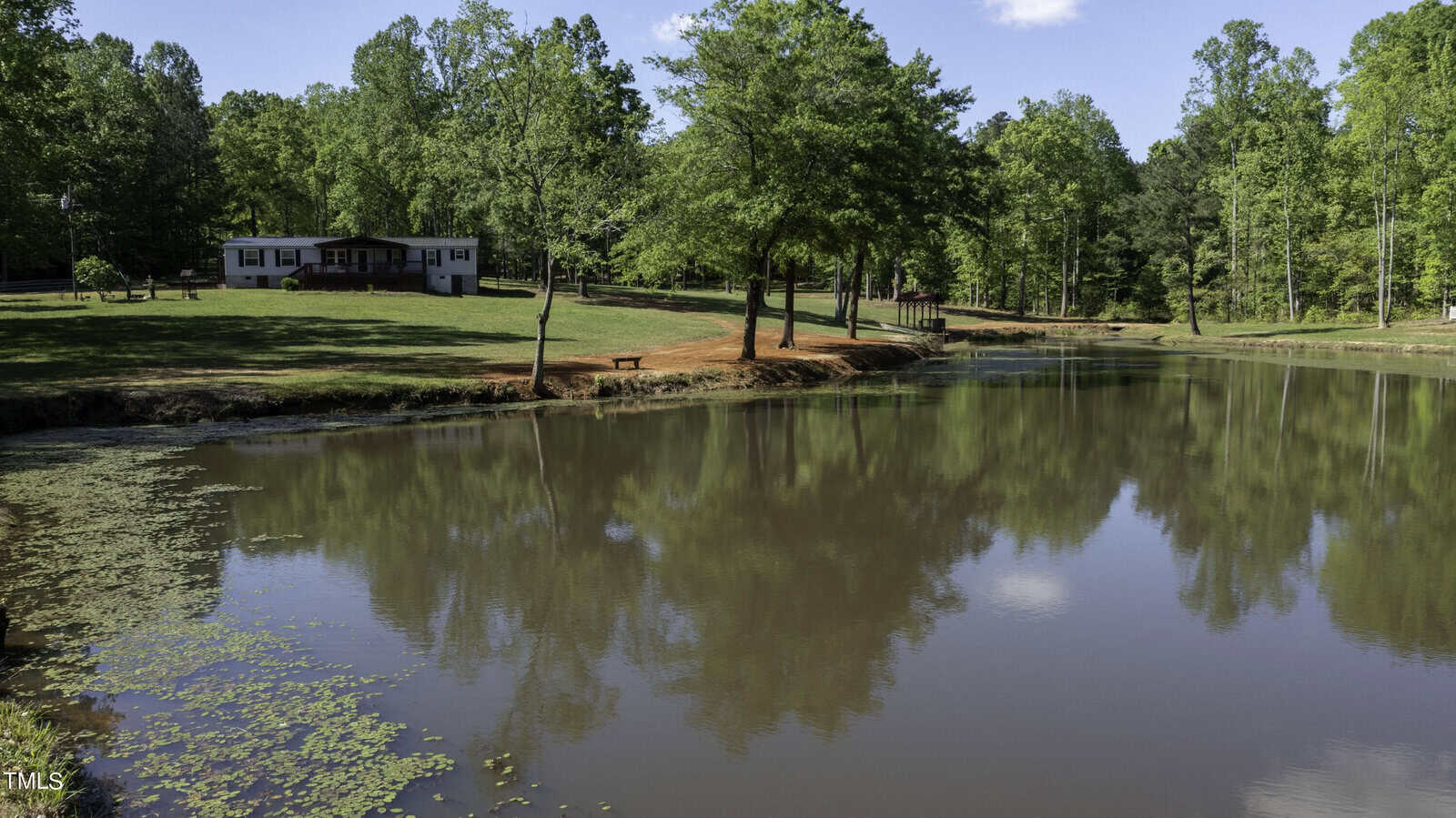 A serene pond reflecting the surrounding trees and blue sky. A house sits on a grassy hill in the background, with lush greenery and a bench along the water's edge.