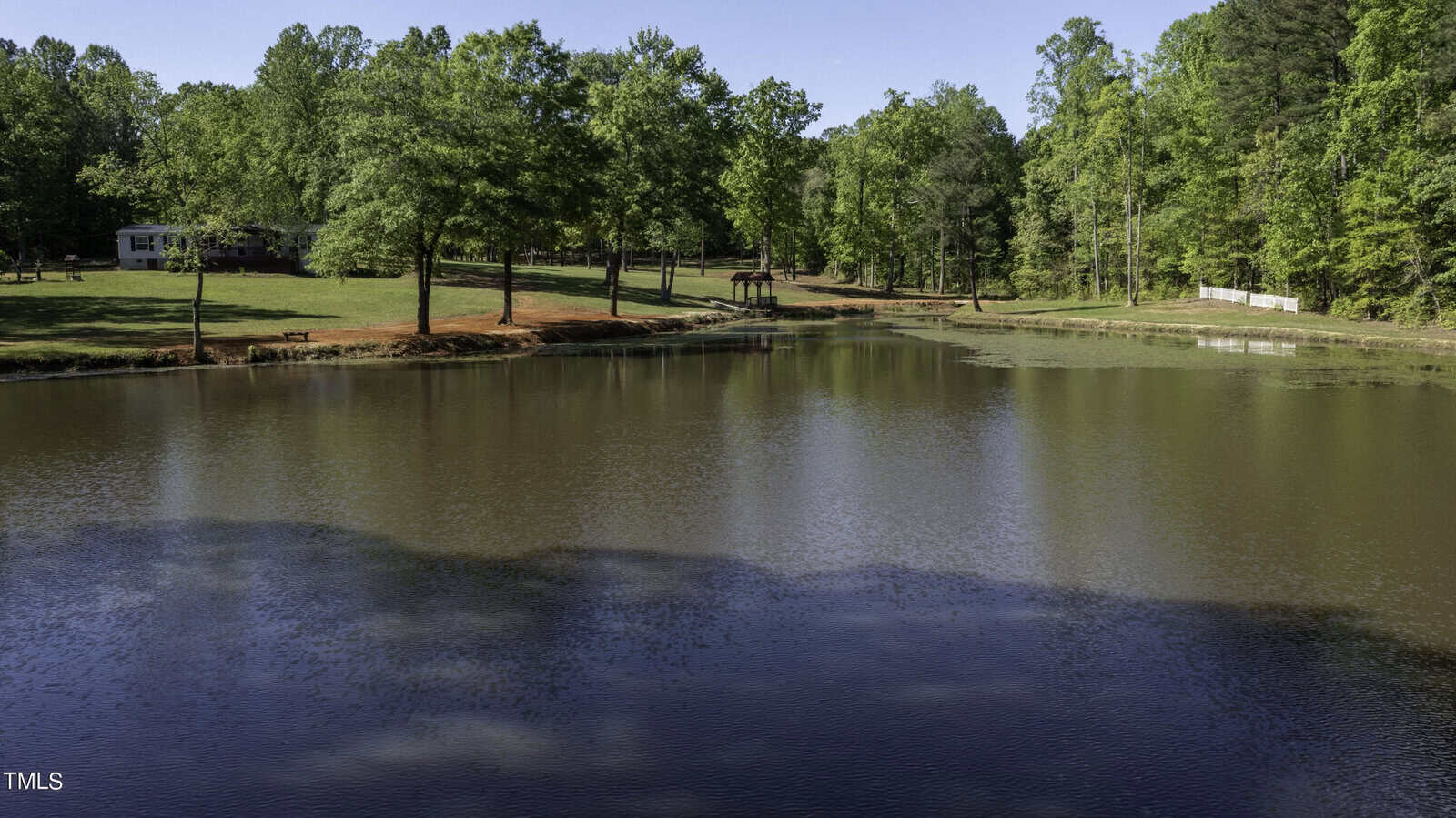 A serene pond surrounded by lush green trees and a grassy lawn. A small house is visible in the background, partially obscured by trees. The water reflects the foliage, creating a peaceful, natural scene under a clear blue sky.