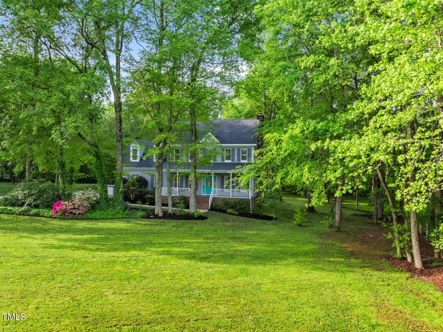 A two-story house with a grey roof is nestled among lush green trees. The home features a white exterior, a spacious porch, and manicured landscaping. A well-maintained lawn stretches across the foreground, with vibrant pink flowers on the left.