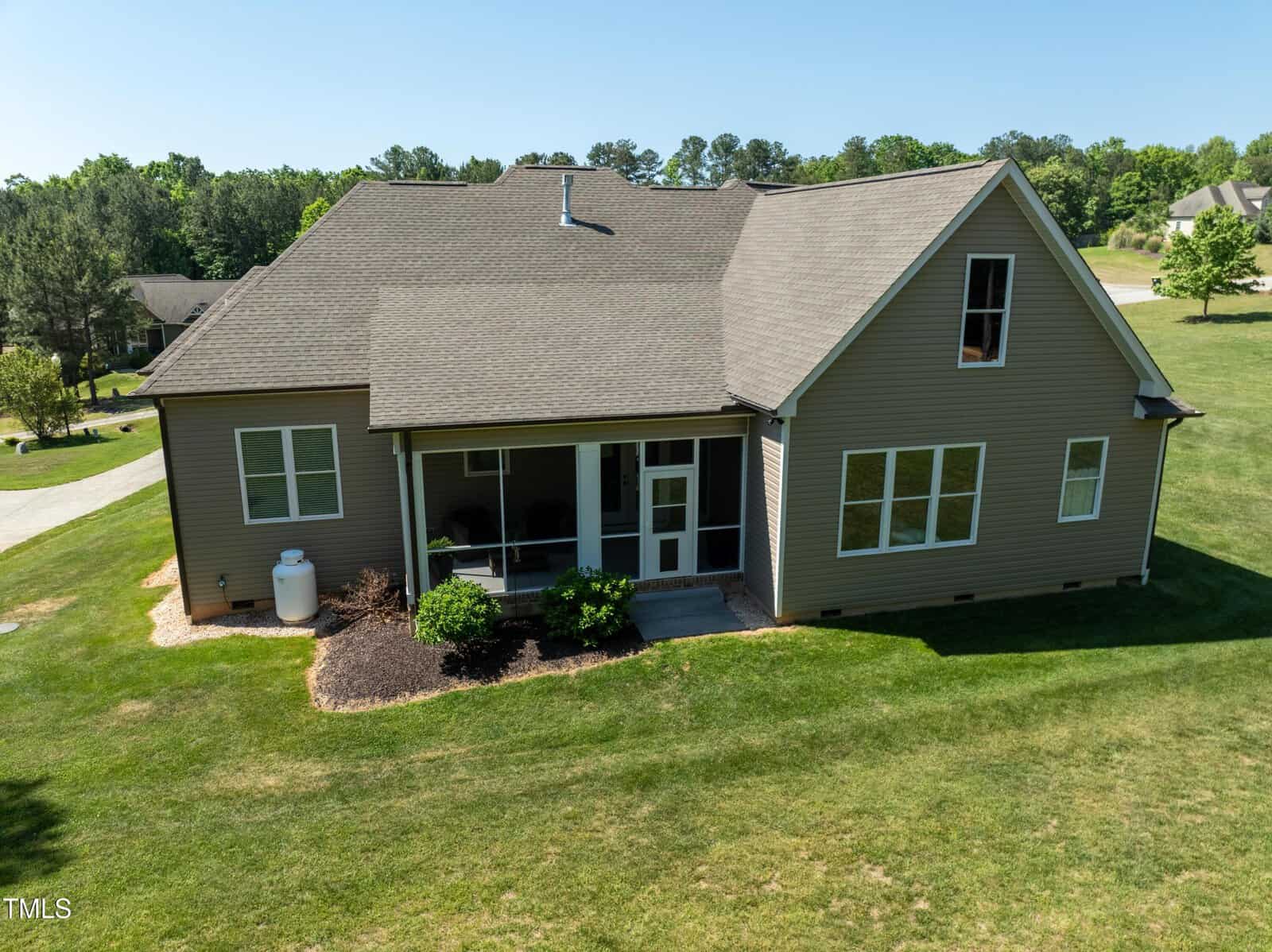 A two-story house with beige siding and a steep roof, featuring a screened porch and several large windows. It's surrounded by a well-kept lawn and trees under a clear blue sky.