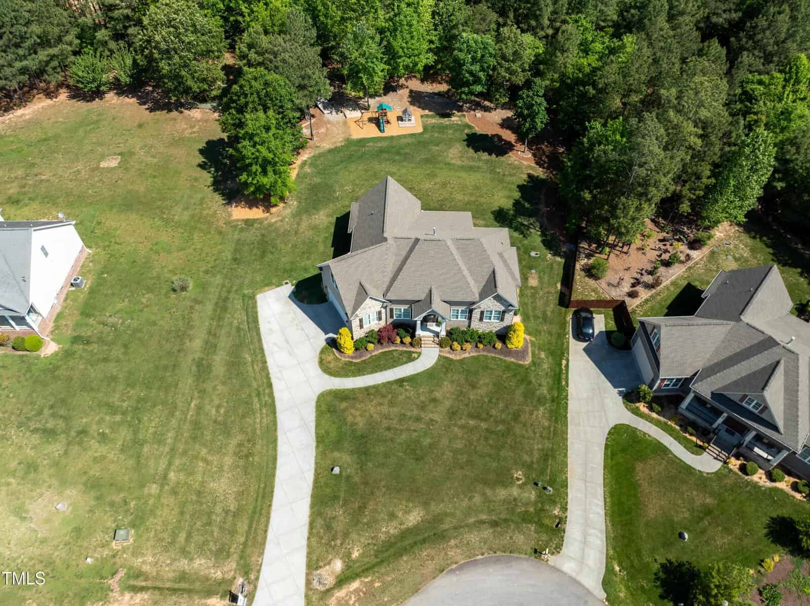 Aerial view of a suburban neighborhood with two houses surrounded by green lawns. A driveway leads to each house. Trees border the backyards, and a small playground is visible in the background.