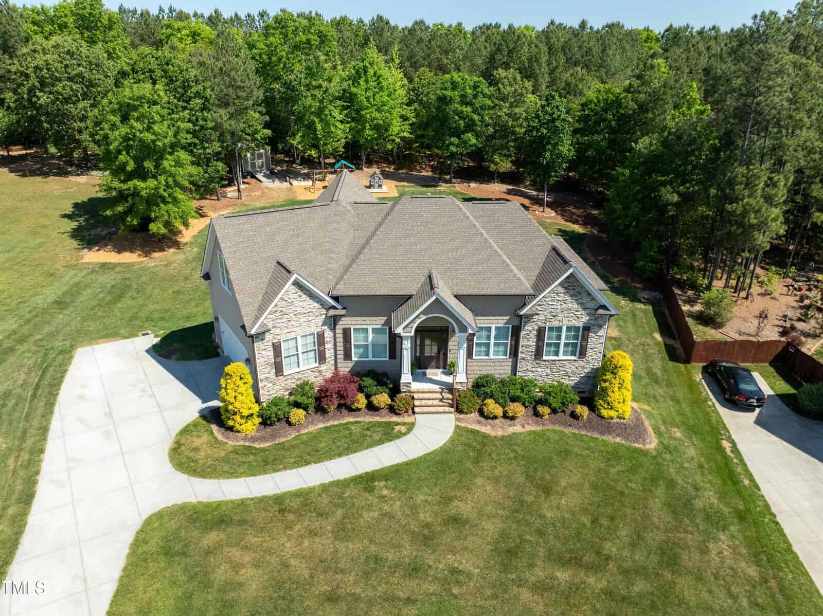 Aerial view of a single-story house with a gray roof and stone facade, surrounded by a well-maintained lawn and landscaped shrubs. A driveway leads to the front of the house. The property is backed by a forested area under a clear blue sky.