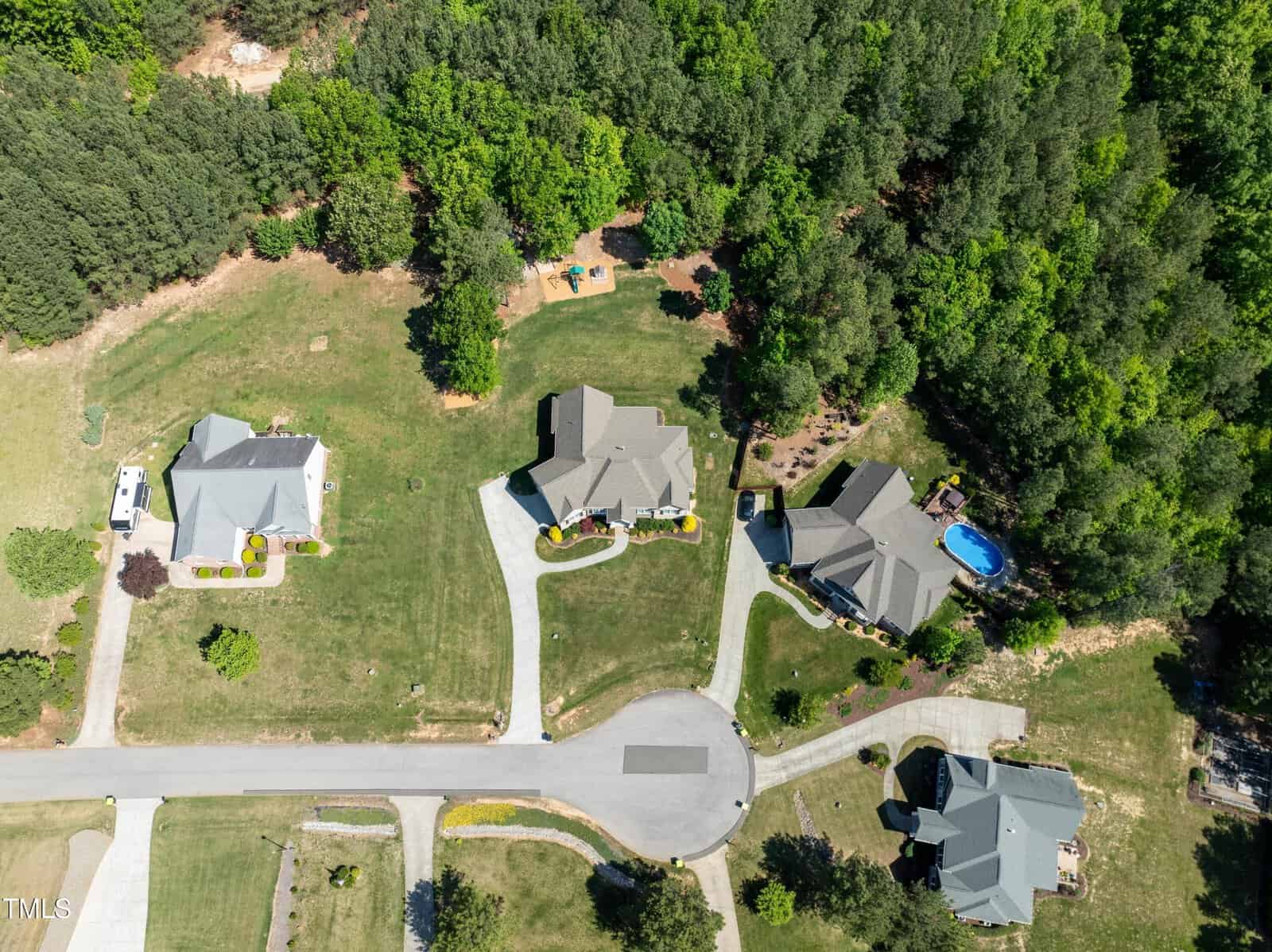 Aerial view of a suburban cul-de-sac surrounded by lush green trees. Four houses with driveways are visible, one with a small blue pool in the backyard. The area appears peaceful and residential.