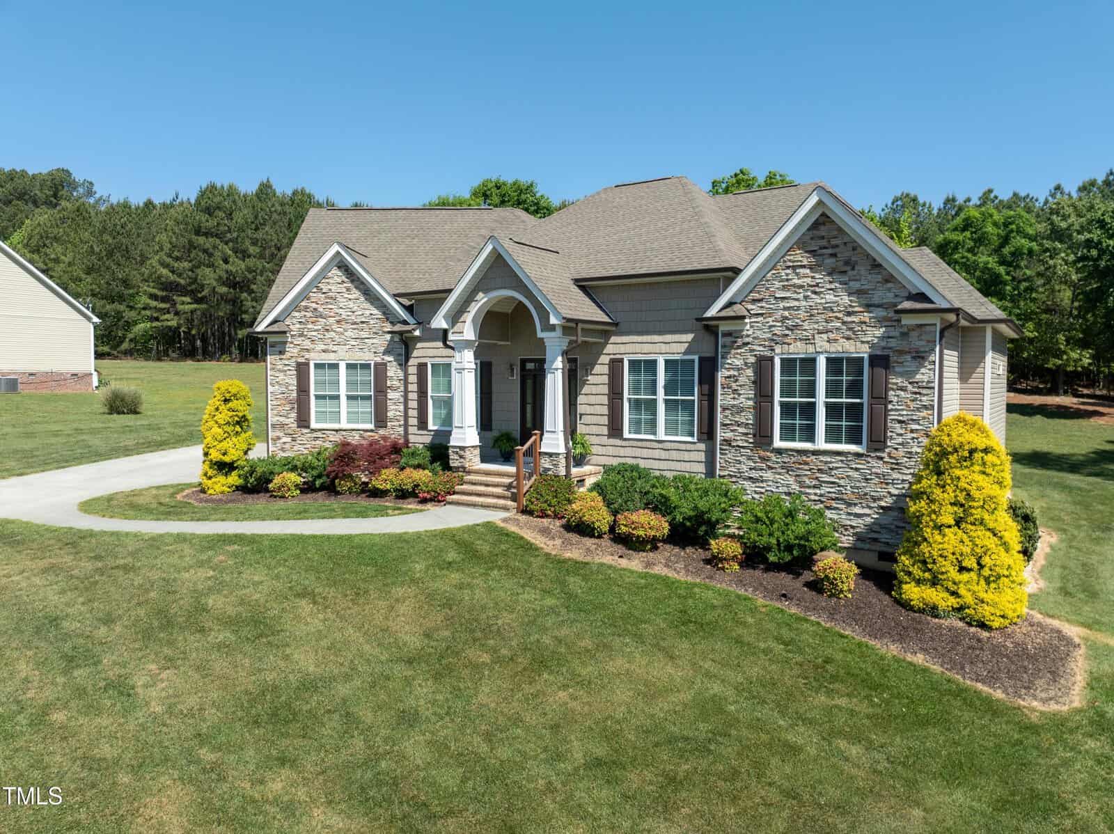 A single-story brick and stone house with a well-maintained lawn, surrounded by greenery and shrubs, under a clear blue sky.