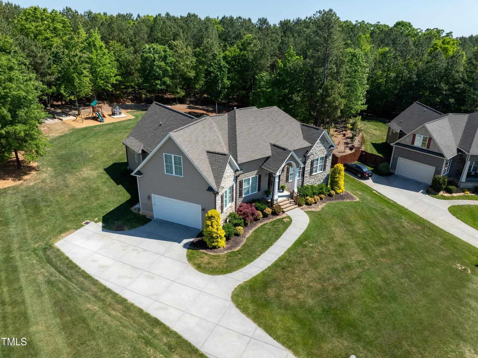 Aerial view of a suburban home with a well-manicured lawn, surrounded by trees. The house has a gable roof, a driveway, and a curved pathway leading to the entrance. A playground is visible in the background.