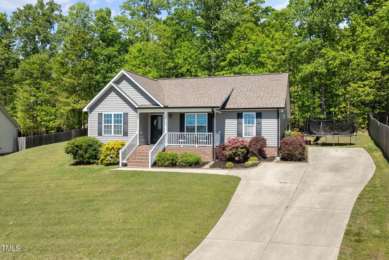 Single-story house with gray siding and brick accents, a front porch, and a well-maintained lawn. A concrete driveway leads to the entrance. A trampoline is visible in the fenced backyard, surrounded by trees.