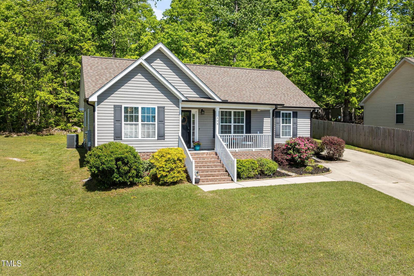 A small, gray house with a front porch and white trim, surrounded by a well-kept lawn and various shrubs. It has a gabled roof and is set against a backdrop of lush green trees. A driveway is on the right.