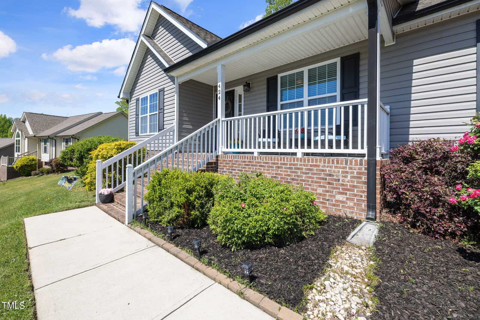 A single-story house with gray siding, a covered porch, and steps leading up. It has a well-maintained front garden with bushes and flowers. The sky is clear and blue.