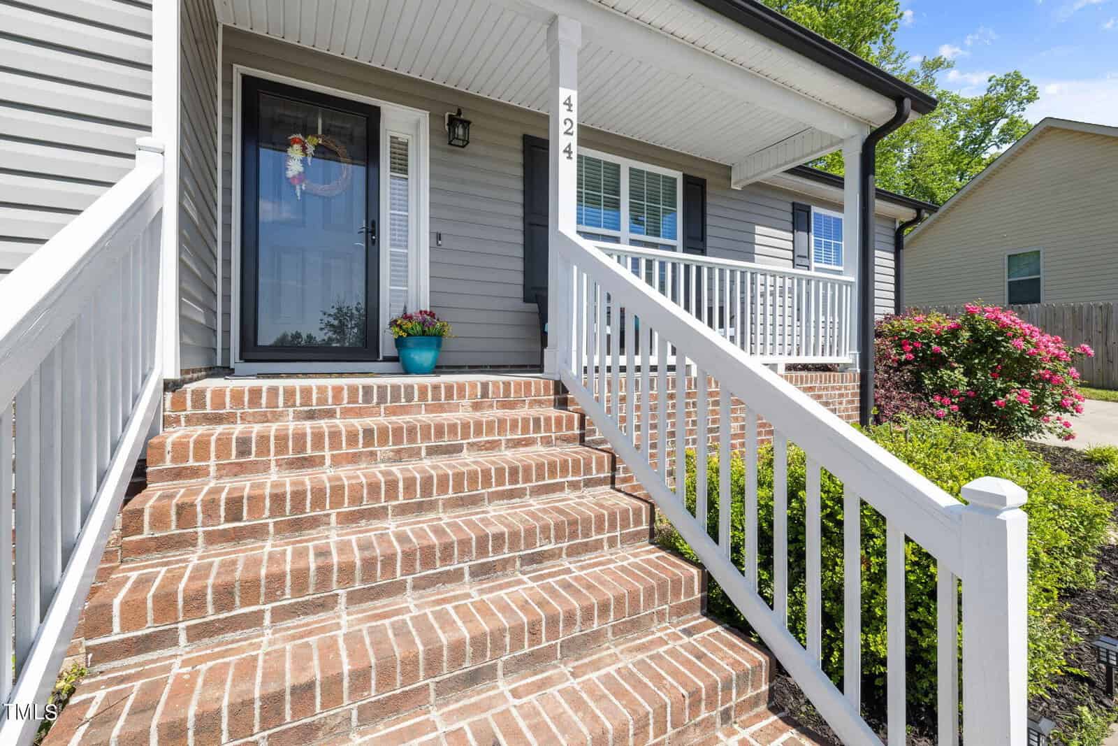 Front porch of a house with brick steps and white railing, leading to a glass-paneled door. A blue flower pot with pink blooms sits beside the door. House number 424 is displayed near the entrance. Bushes with flowers are visible to the right.