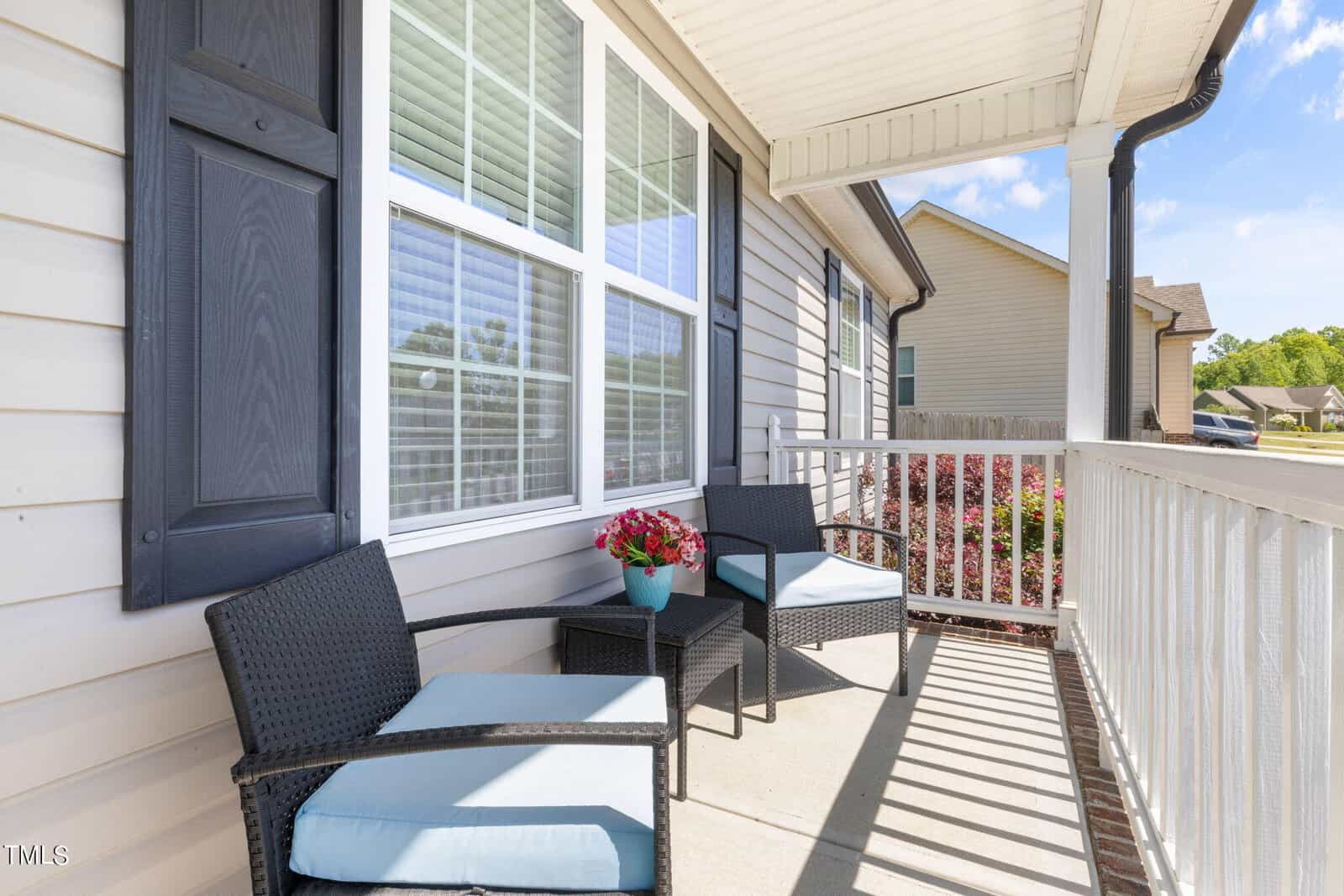 A small porch featuring two black wicker chairs with light blue cushions, a matching side table holding a potted pink flower, all against a white siding house with a large window. A white railing encloses the porch.