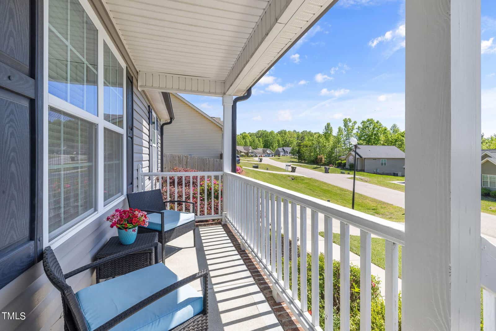 A cozy porch with two black wicker chairs and blue cushions is adorned with pink flowers on a small table. The porch overlooks a quiet suburban street with green lawns and houses under a clear blue sky.