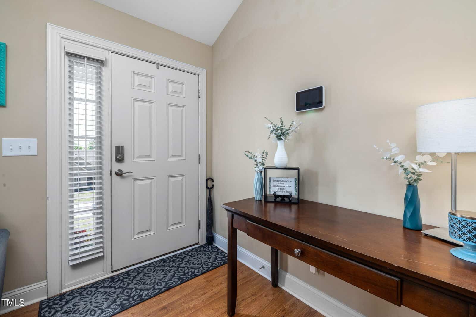 A cozy entryway with a wooden table holding white floral arrangements in vases and a small framed sign. A table lamp with a blue base is on the right. The door has a glass panel with blinds. A blue patterned rug lies on the wooden floor.