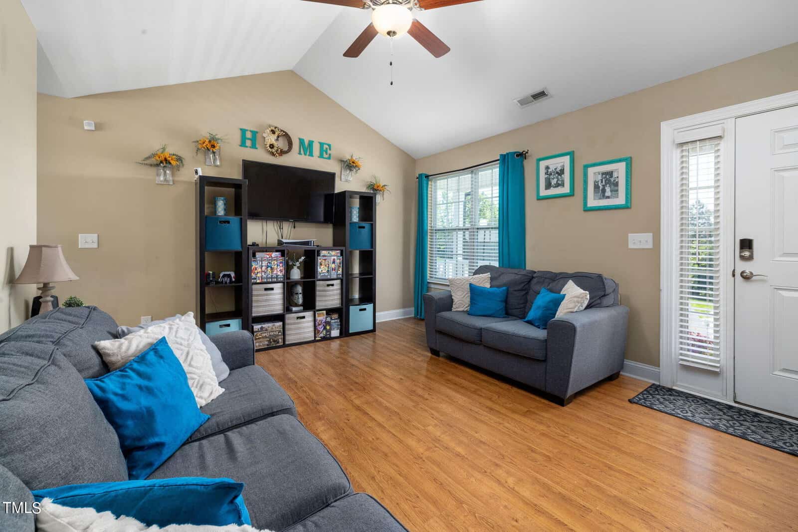 A cozy living room with a high ceiling, featuring two gray sofas with blue and white cushions. A TV and shelving unit hold decorative items under a "HOME" sign. The room has wood flooring, and natural light comes through a window and glass-paneled door.