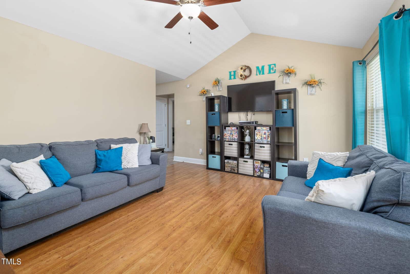 A cozy living room with two blue sofas, white and teal throw pillows, and a wooden floor. A large shelving unit holds a TV and decorative items. Blue curtains flank the window on the right, and a ceiling fan is above. The wall spells "HOME" with decor.