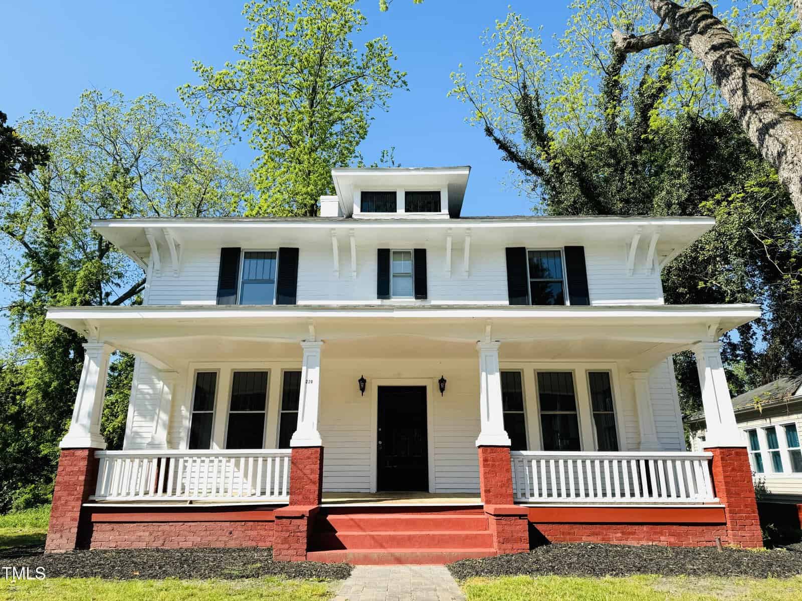 A large, two-story white traditional house at 220 Charles Street, Henderson NC with a broad front porch, flanked by tall trees under a clear blue sky.