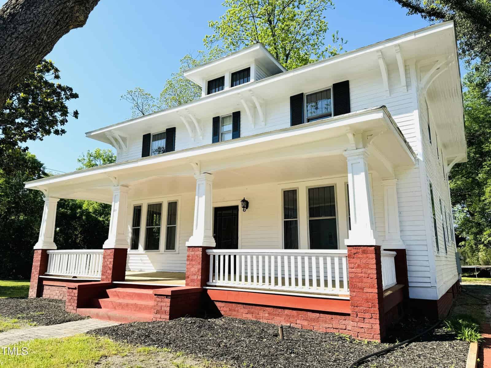 A two-story white house with a large covered porch and red brick foundations, surrounded by greenery under a clear blue sky.