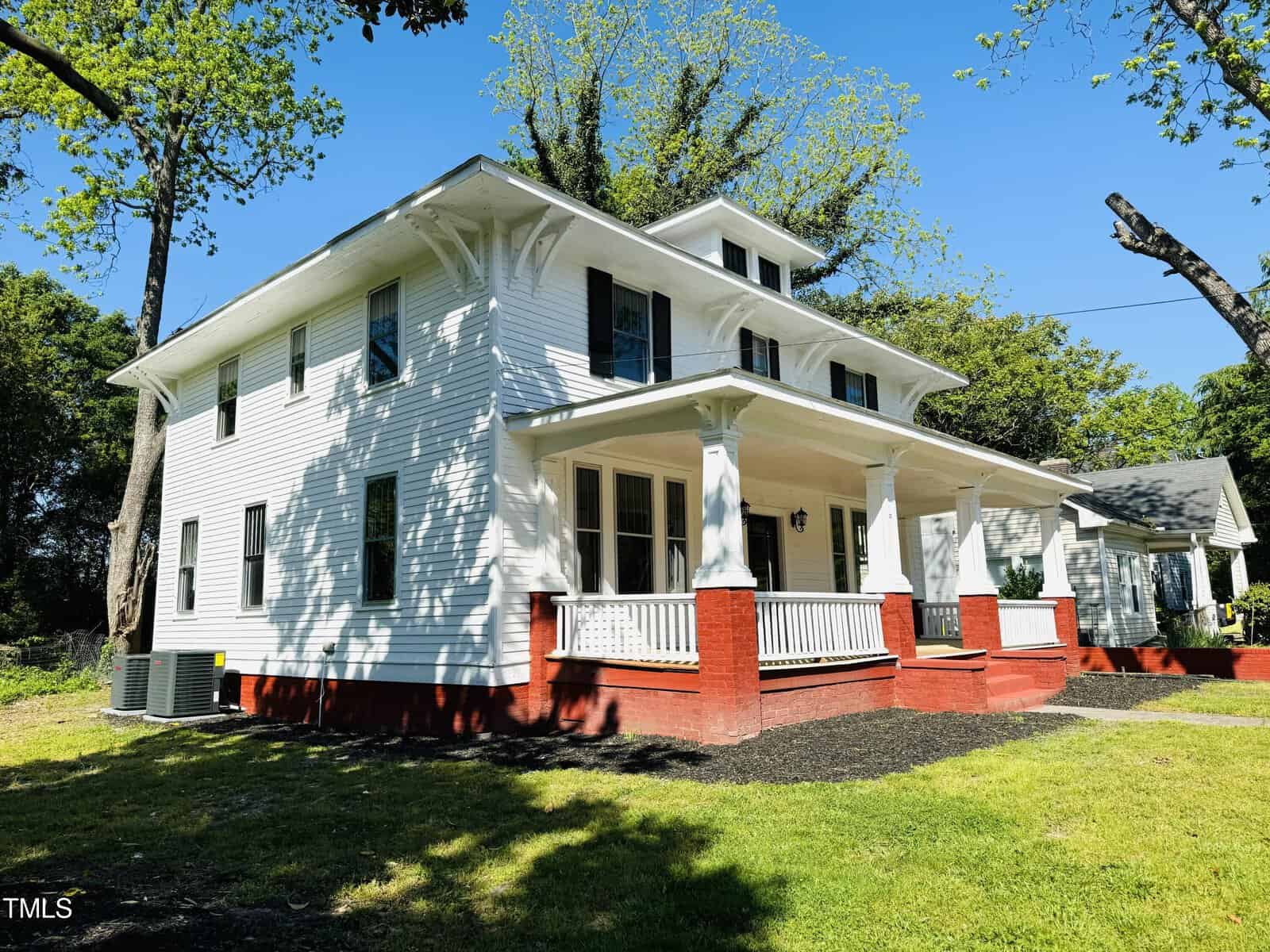 A traditional two-story white house with a wide front porch, red steps, and surrounded by lush greenery under a clear blue sky.
