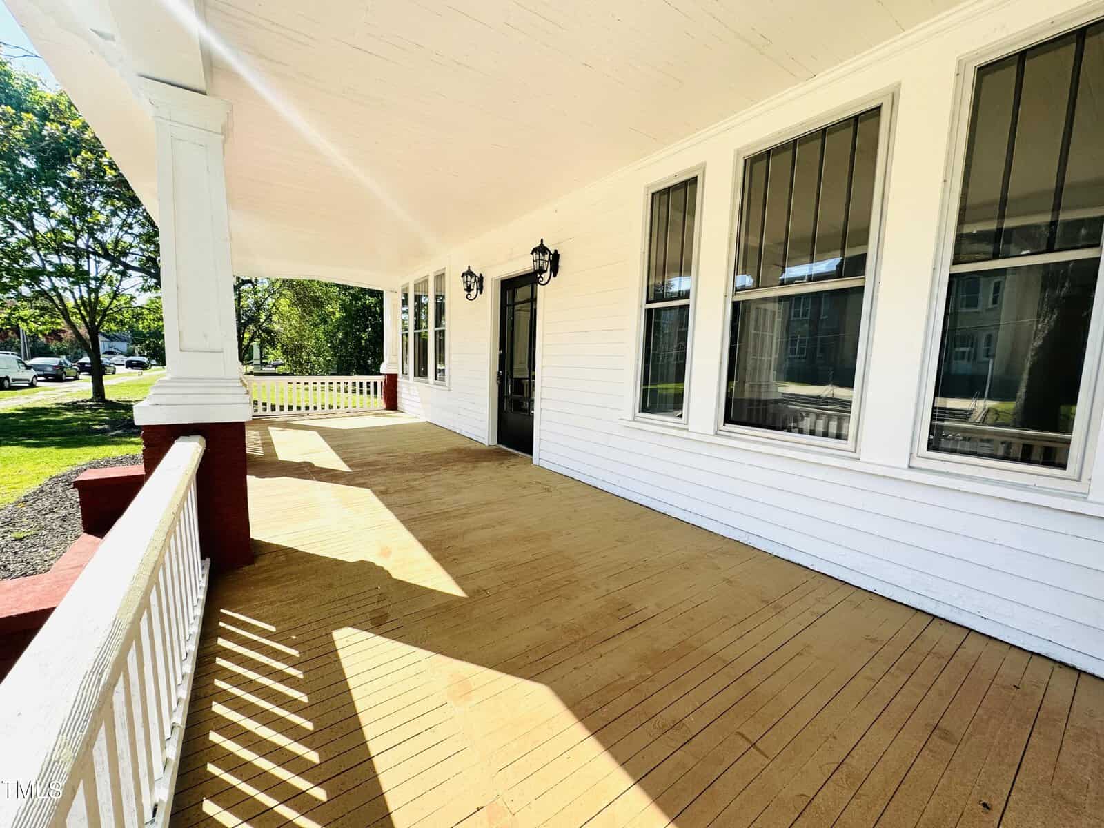 A spacious wooden porch of a white house with white columns, windows covered with blinds, a black lamp, and a white railing, overlooking a grassy area with trees.