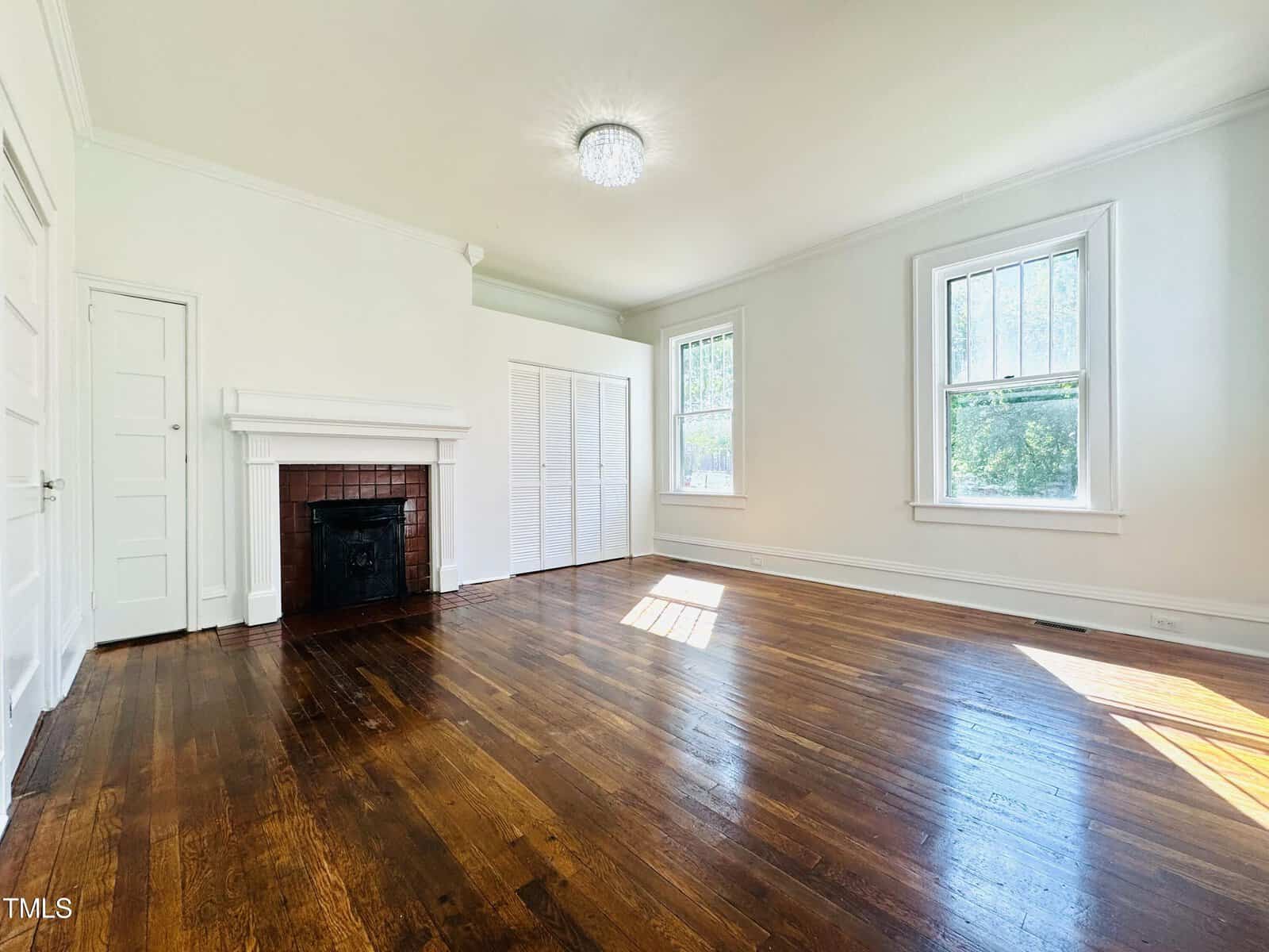 Bright, spacious room featuring polished hardwood floors, a fireplace, large windows, and a white door, set against white walls, bathed in natural light.
