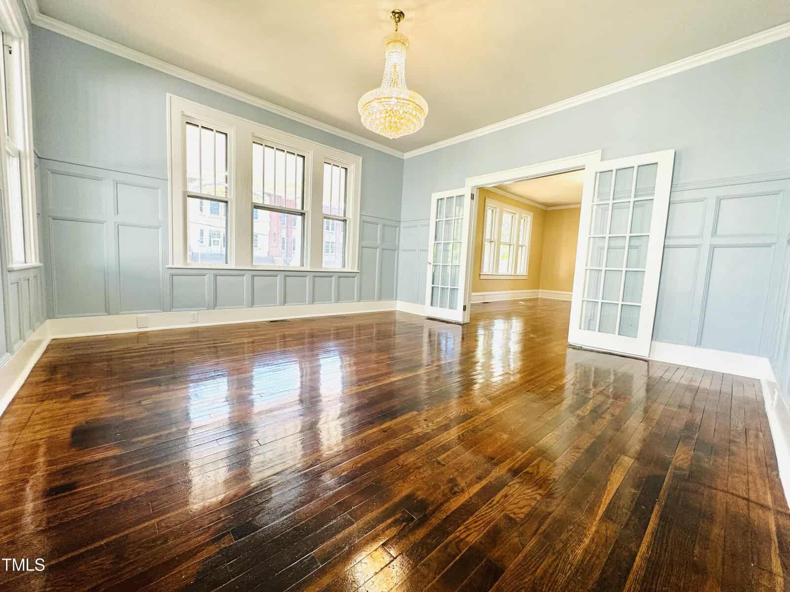 An elegant empty room with polished hardwood floors, light blue walls with white wainscoting, large windows, and open french doors leading to another room. a crystal chandelier hangs from the ceiling.