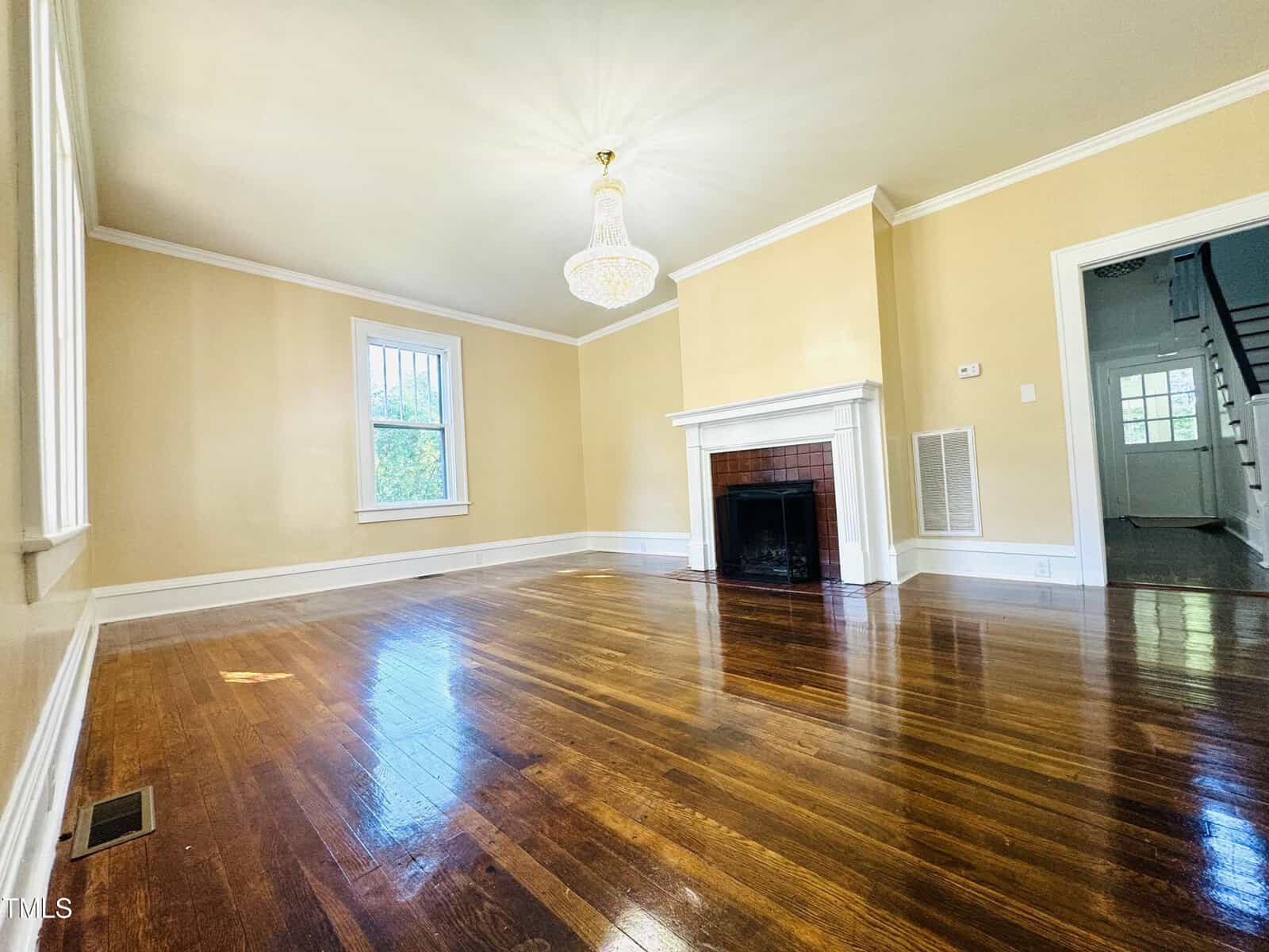 Spacious living room with glossy hardwood floors, a traditional fireplace, light yellow walls, and two large windows. an elegant chandelier hangs from the ceiling.