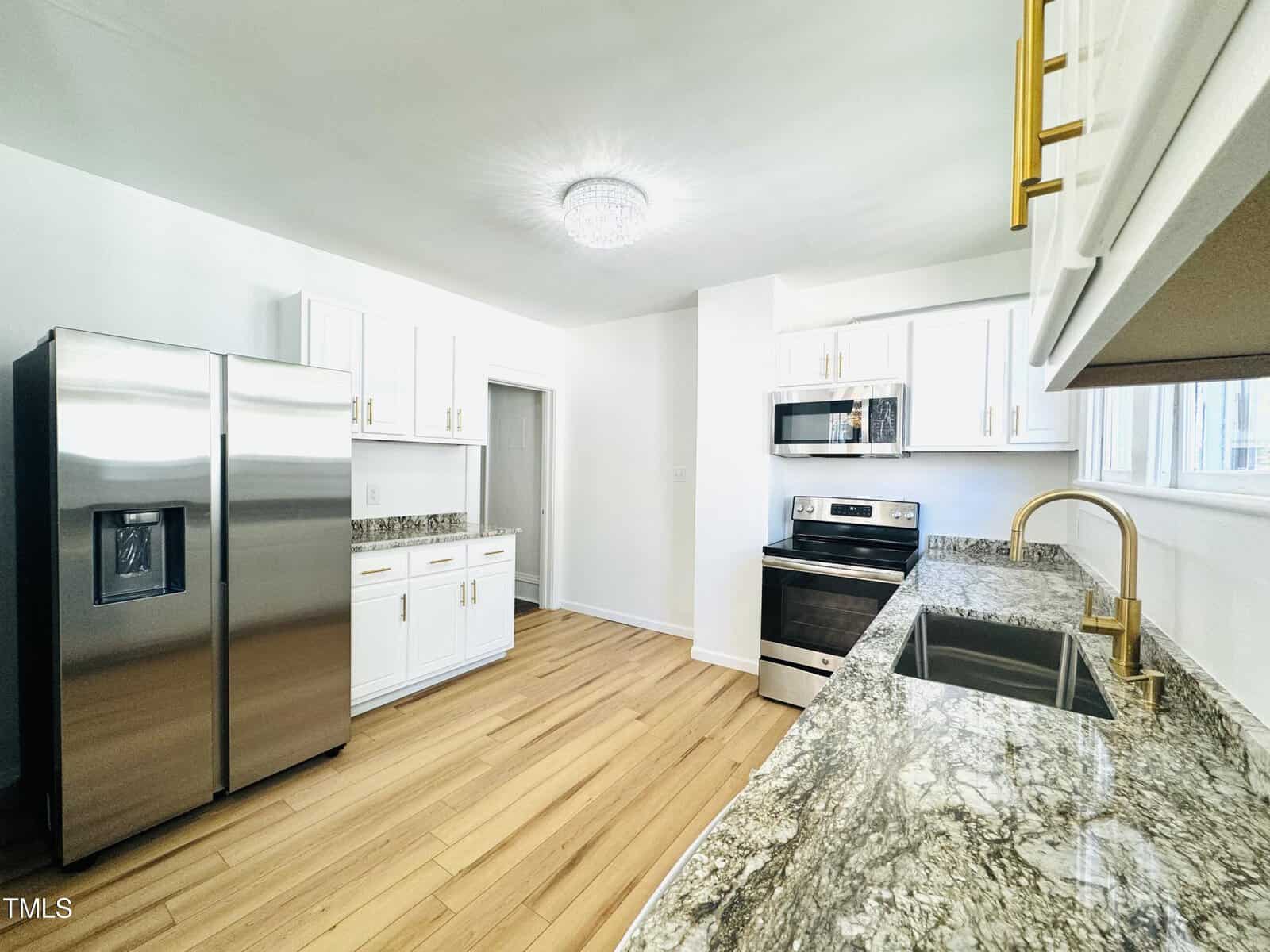 A bright, modern kitchen featuring white cabinets, stainless steel appliances, a marble countertop, and light wooden floors.