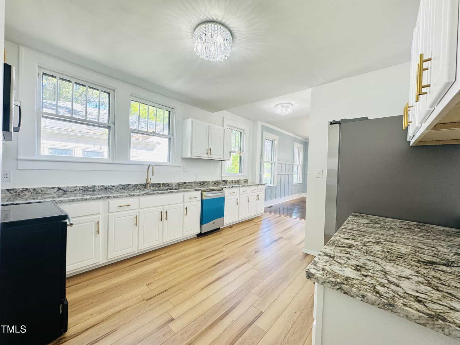 A bright, modern kitchen with white cabinetry and granite countertops, featuring stainless steel appliances, wooden floors, and large windows letting in natural light.
