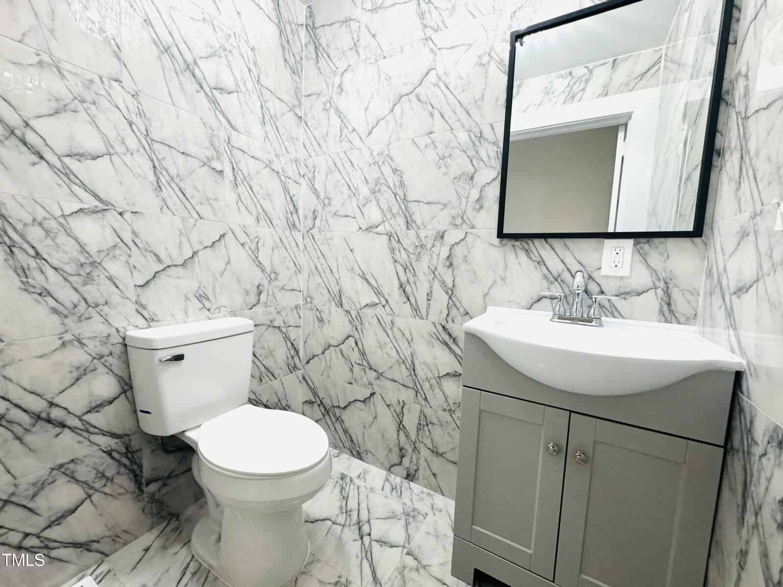 A modern bathroom featuring walls and floor fully clad in white and grey marble. it includes a white toilet, grey vanity with a sink, and a rectangular mirror.