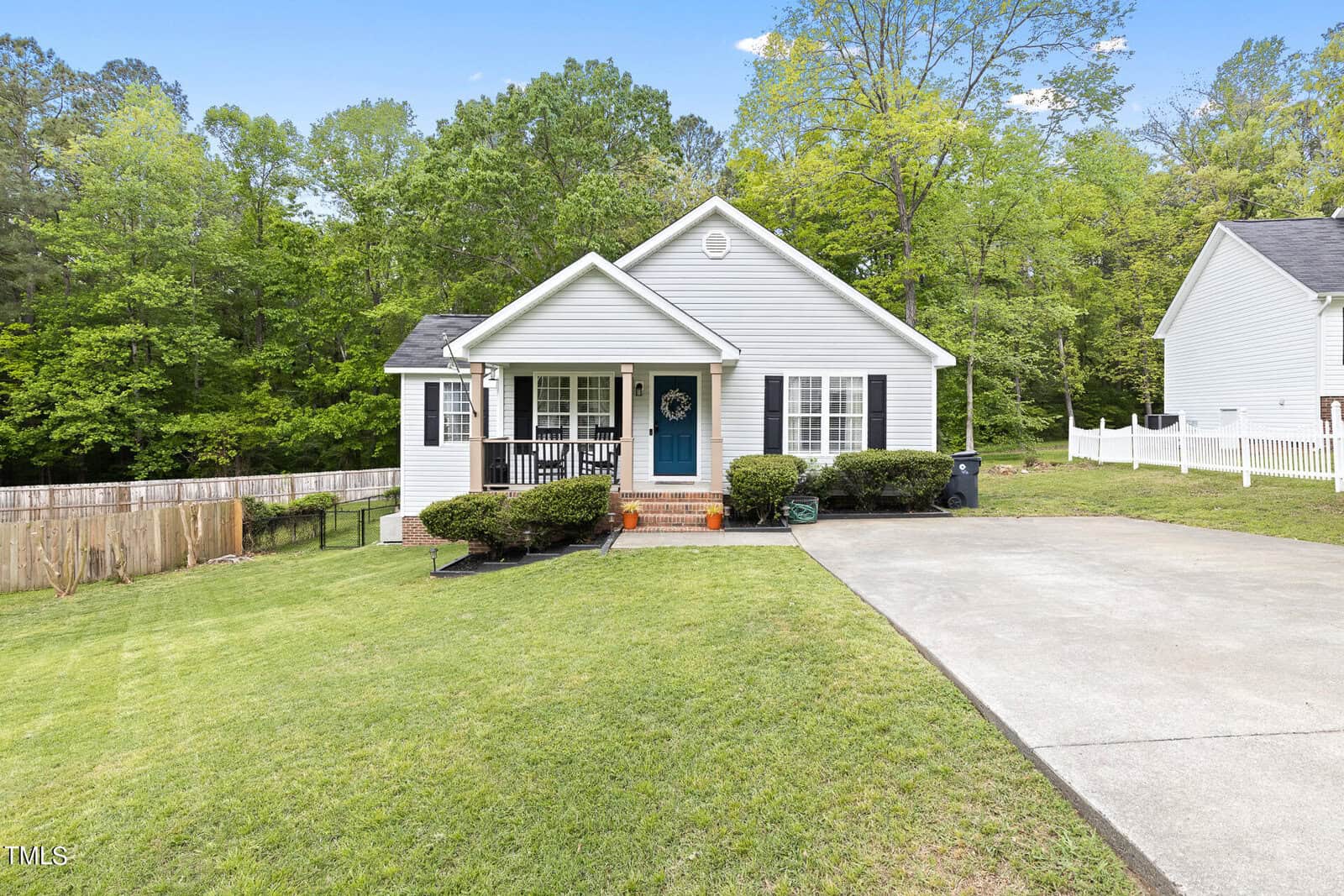 A charming one-story house at 107 Margaret Drive in Creedmoor boasts a light gray exterior and a bright blue front door, surrounded by a neatly manicured lawn. A concrete driveway lies to the right, and on the left is a wooden fence, framed by lush green trees in the background.