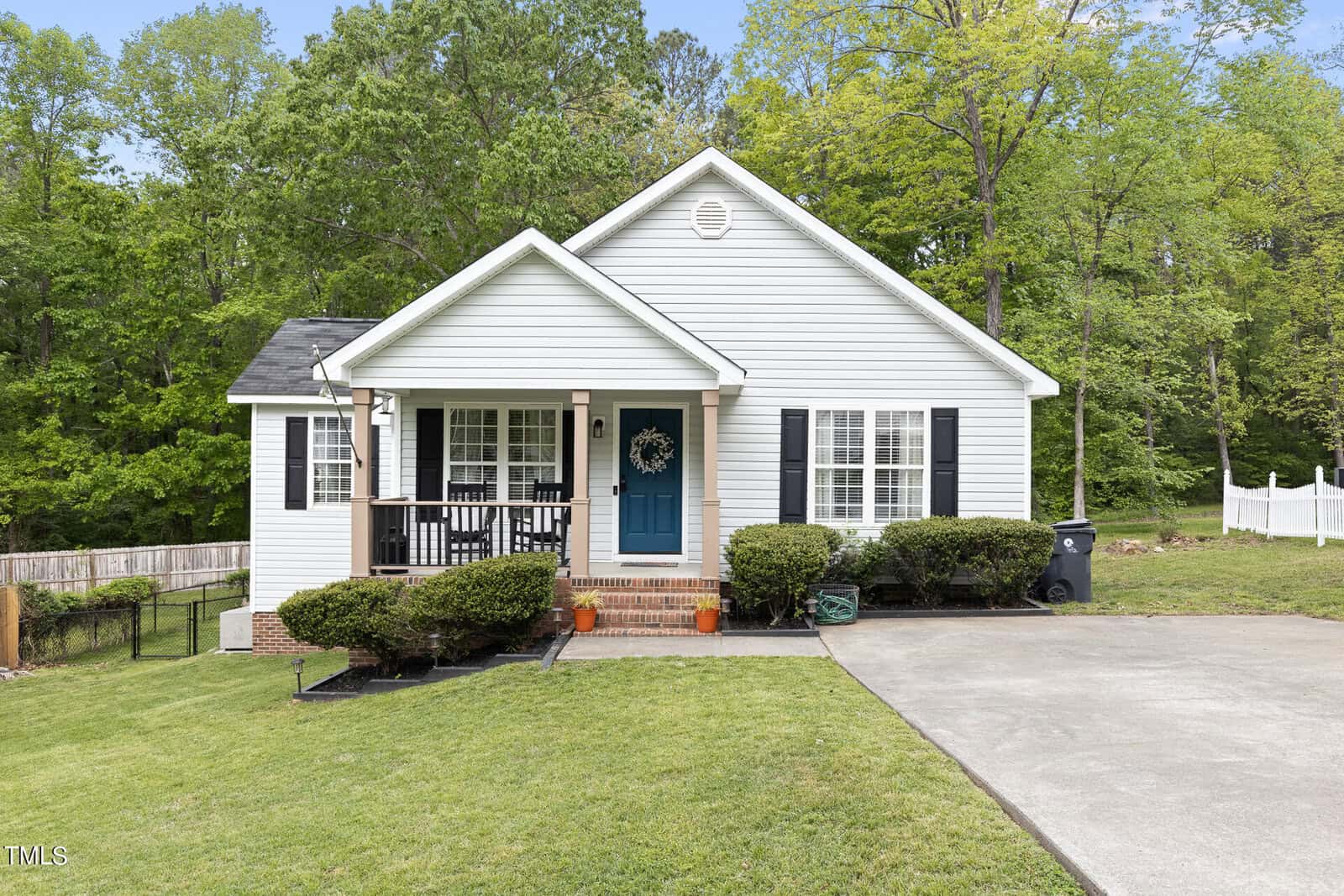 A single-story white house with blue shutters and a teal door, nestled among lush green trees. A small front porch features a decorative wreath. The lawn is neatly mowed, and a paved driveway is visible to the right.