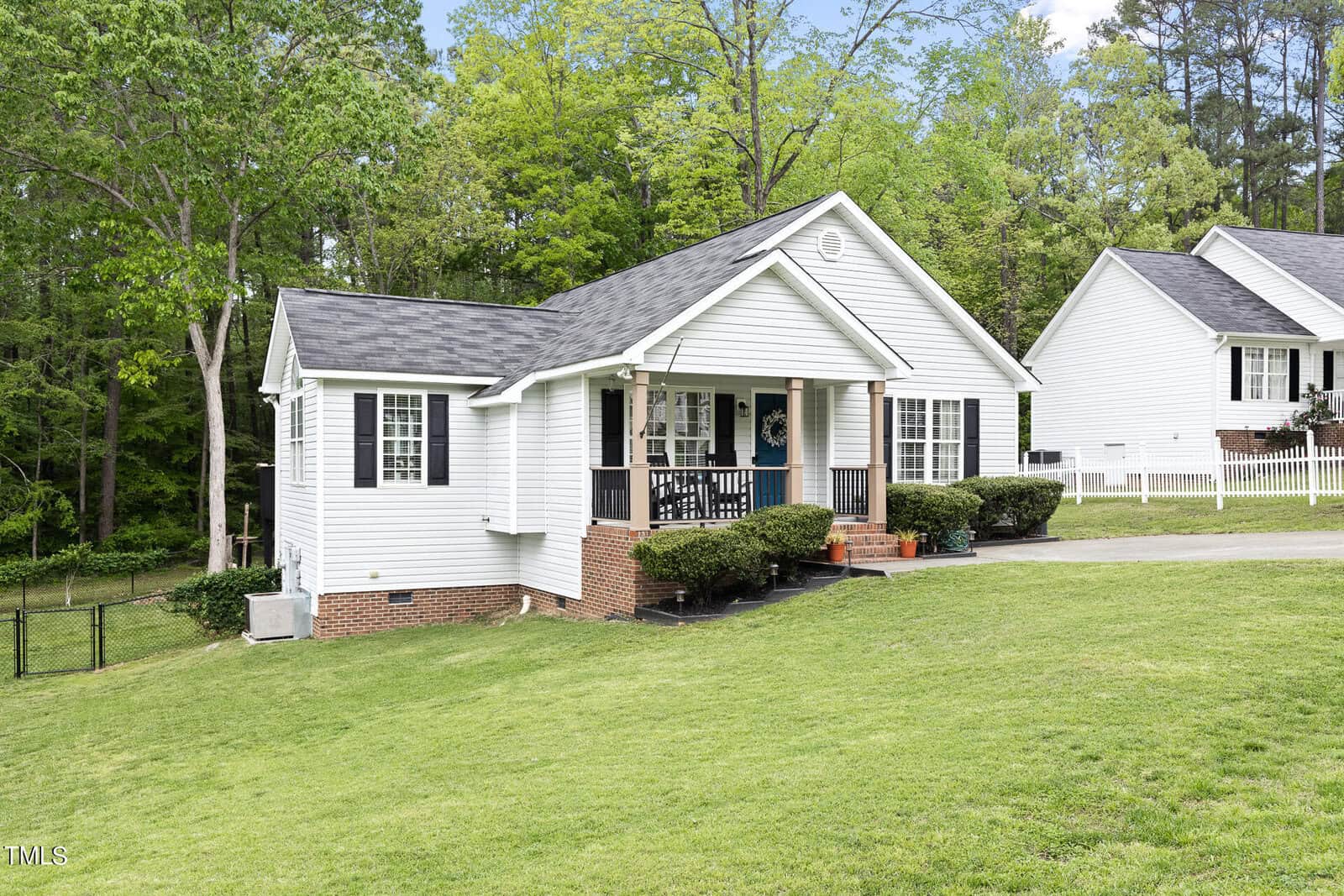 A white, single-story house with a black shingled roof surrounded by green grass and trees. The house has a porch with steps, railings, and bushes in front. A neighboring house and a fence are visible in the background.