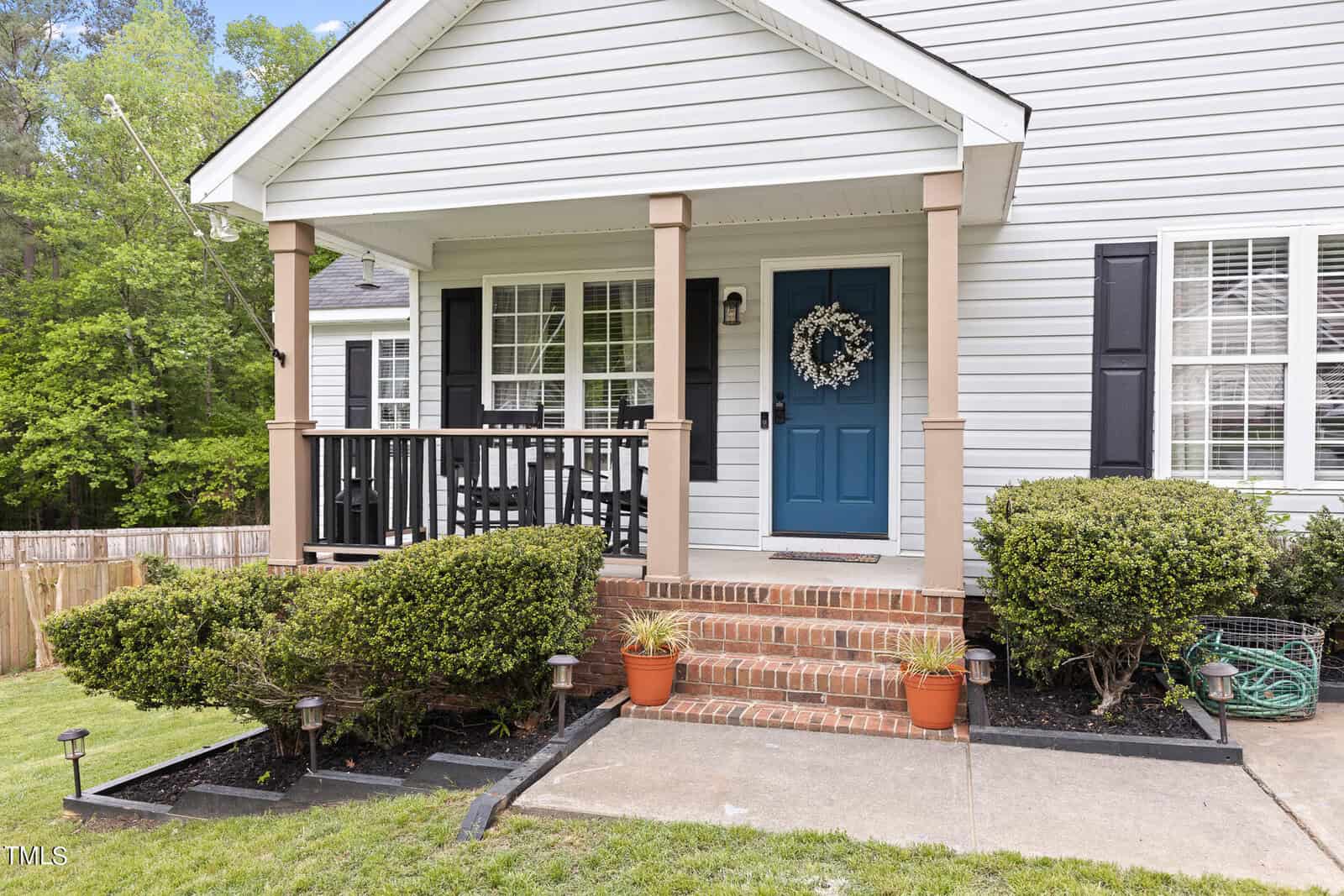 A small house with white siding and a blue front door adorned with a wreath. The porch has black railings and is bordered by shrubs. A few potted plants line the brick steps leading to the entrance. The yard has a concrete walkway.