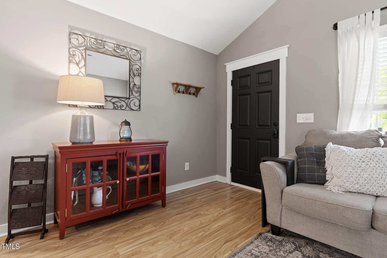 A cozy living room with a gray couch and decorative pillows, a red cabinet with glass doors, and a decorative mirror above. A black door is on the right, and a small wall shelf holds two small items. The floor is light wood, and a lamp sits on the cabinet.
