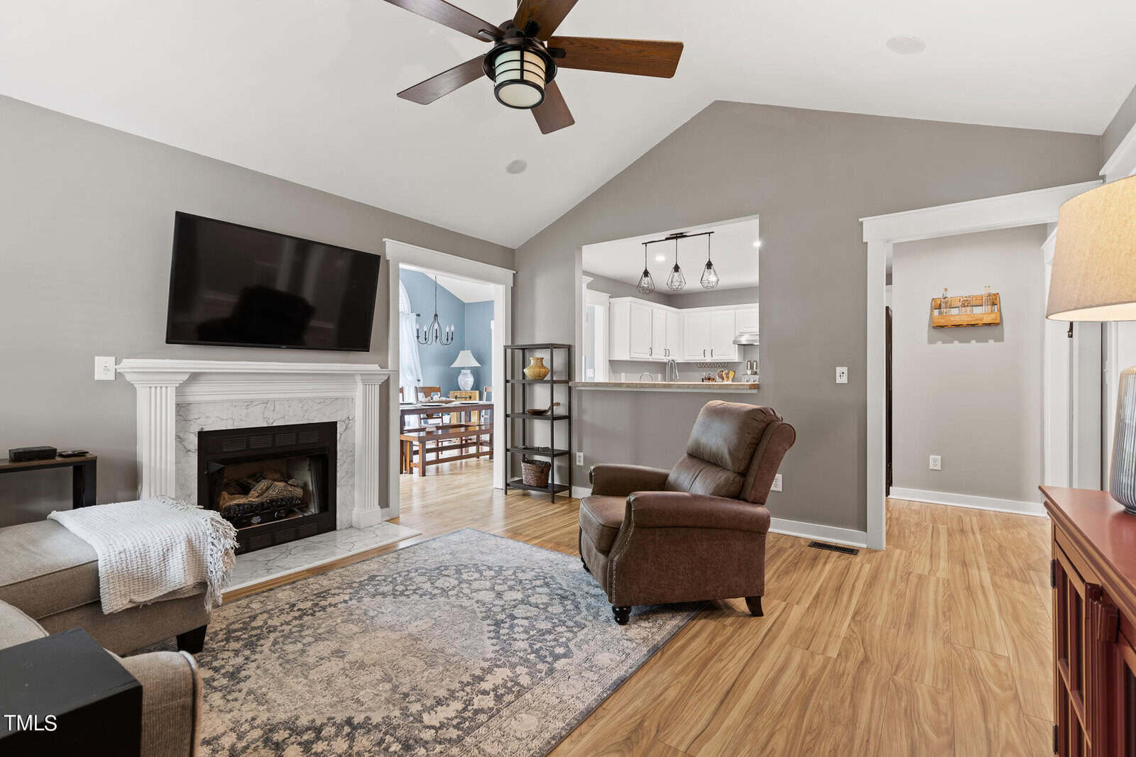 A cozy living room with a brown recliner, flat-screen TV above a white fireplace, and a ceiling fan. The room features wooden floors, gray walls, and a patterned rug. A view into the dining area and kitchen is visible through a large doorway.