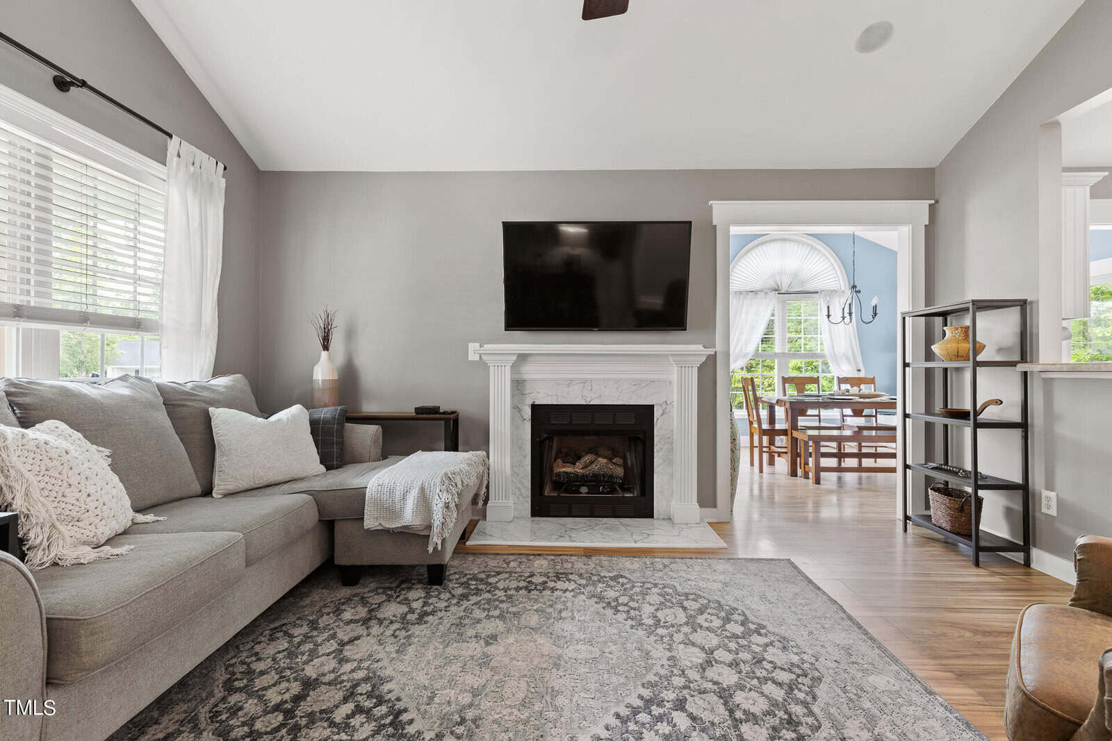 A cozy living room with a gray sectional sofa, a patterned area rug, and a fireplace topped with a flat-screen TV. The room leads to a dining area with a table and chairs, visible through a wide doorway. Natural light fills the space.