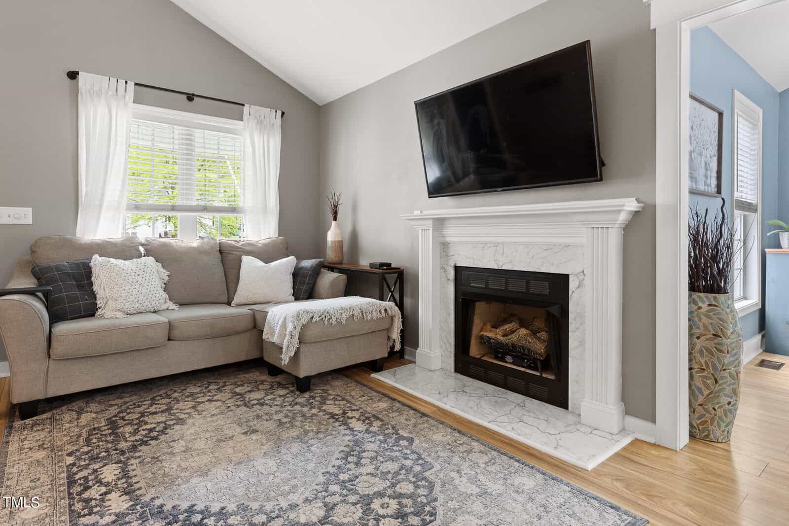 Cozy living room with a gray sofa, patterned rug, and pillows. A TV is mounted above a white fireplace, and a window with white curtains lets in natural light. A small table with a vase is beside the sofa. Walls are painted in neutral tones.