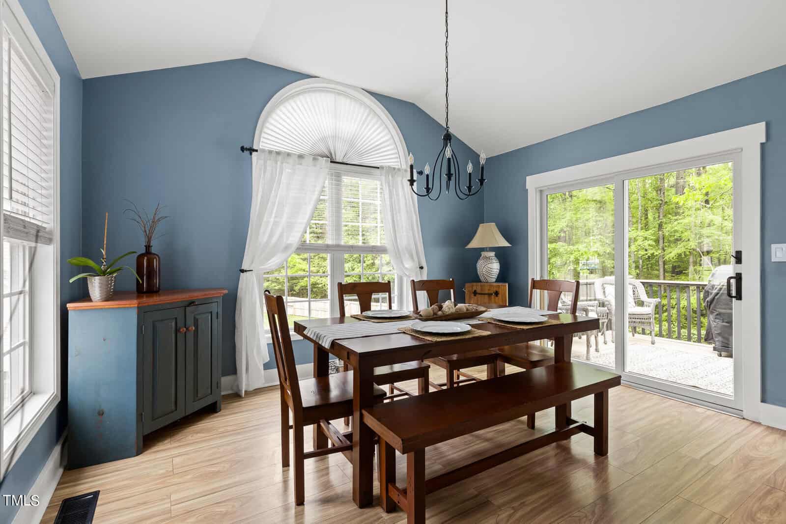 A dining room with a wooden table and bench, surrounded by chairs. Large windows with white curtains let in natural light. The walls are painted blue. A glass door opens to a patio, and there's a chandelier overhead. A sideboard holds decorative items.