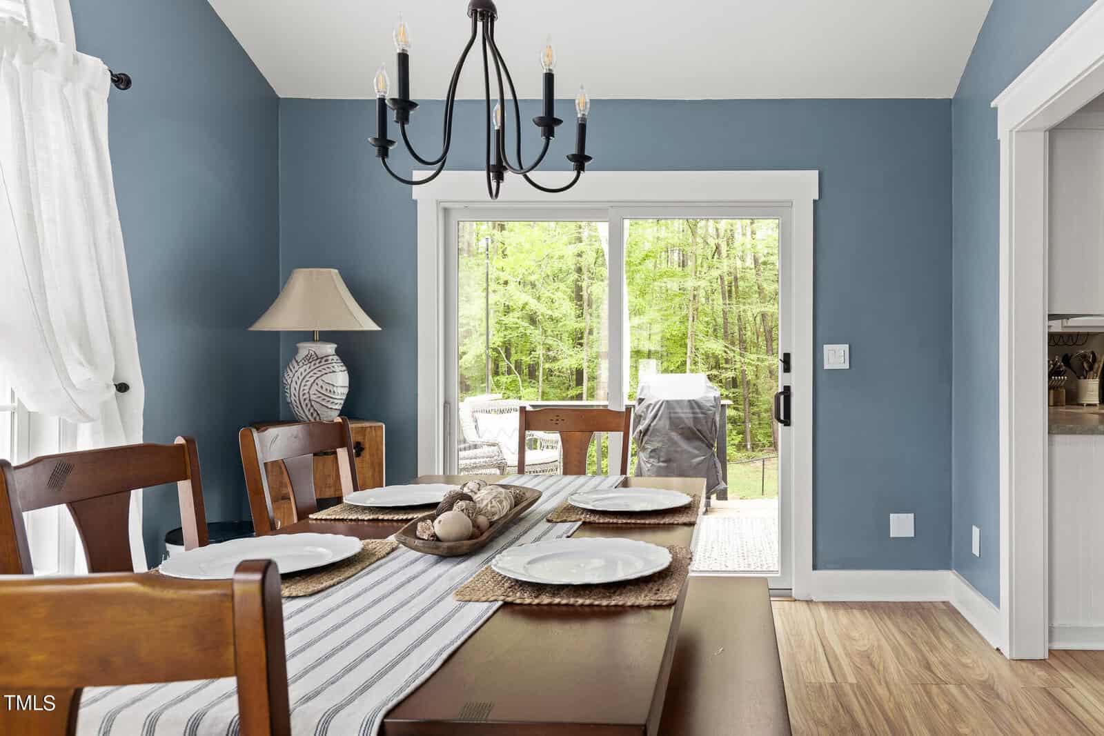 Dining room with a wooden table set with plates and a striped runner. A black chandelier hangs above. A sliding glass door reveals a view of a green forest and outdoor grill. Blue walls and a white curtain accent the space.