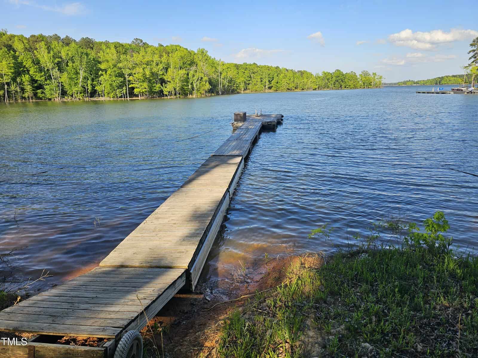 A long wooden pier extends into a serene lake, surrounded by lush green forests under a clear blue sky.