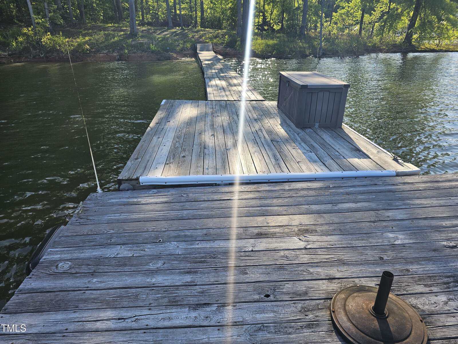 A wooden dock extends into a tranquil lake, surrounded by lush green trees, with a fishing rod resting along one side and a small metal object on the foreground edge. bright sunlight casts long shadows on the dock.
