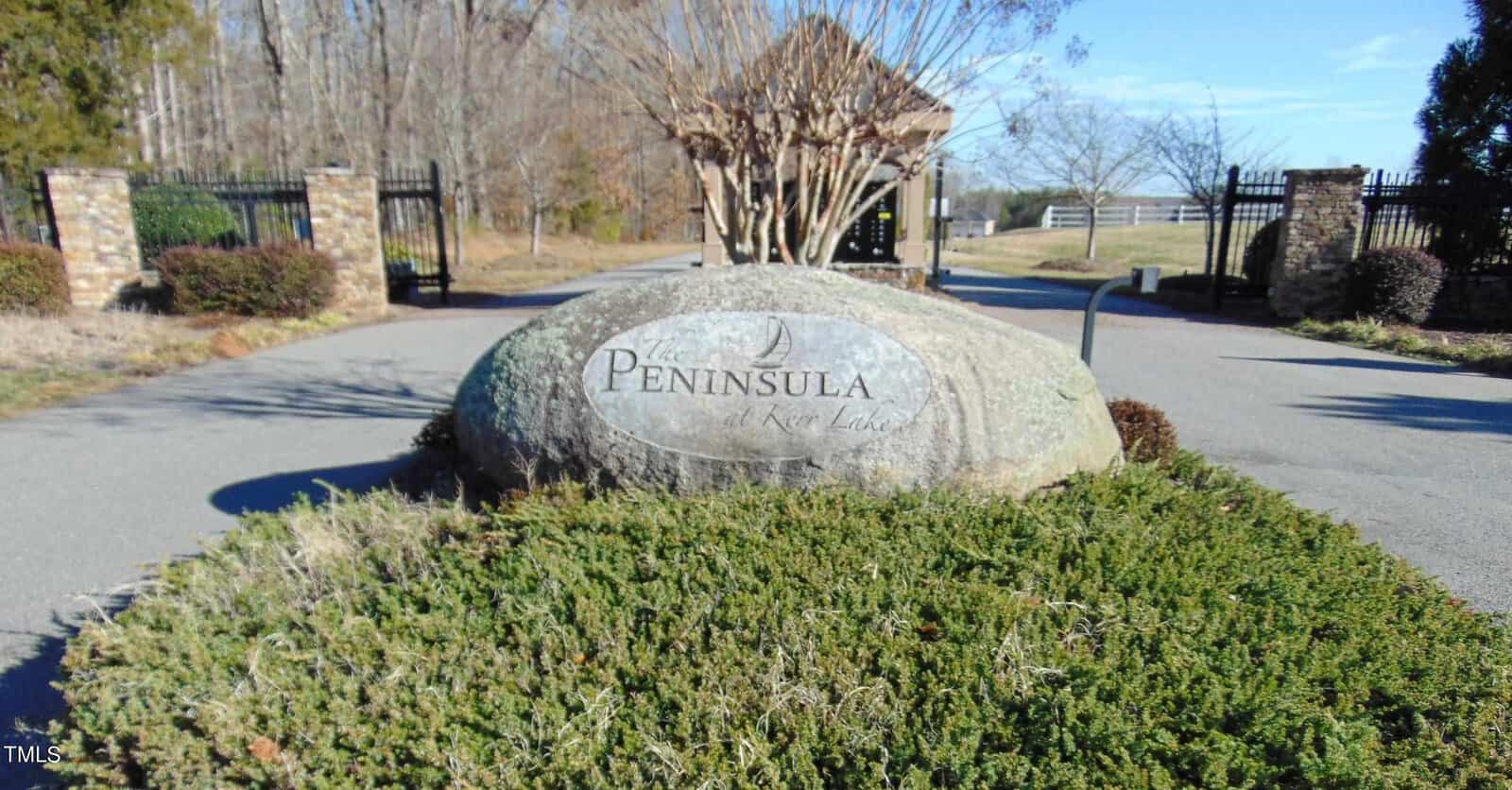 A large engraved stone reading "the peninsula at kerr lake" is nestled among shrubbery at the entrance gate of a community, with an open iron gate and a clear blue sky in the background.