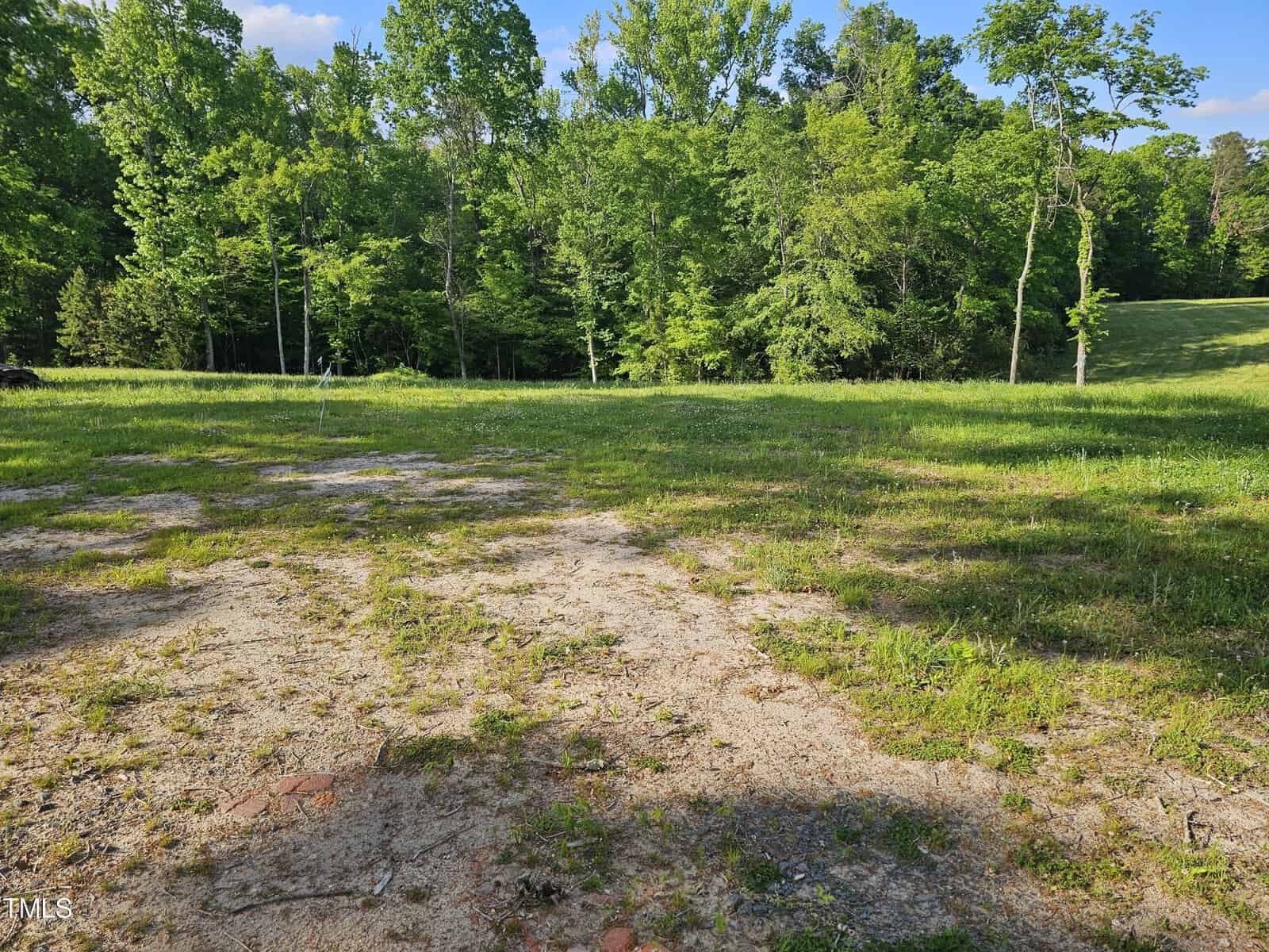 A scenic view of a grassy field transitioning into a wooded area under a clear blue sky. the foreground shows patches of bare earth and sparse grass.