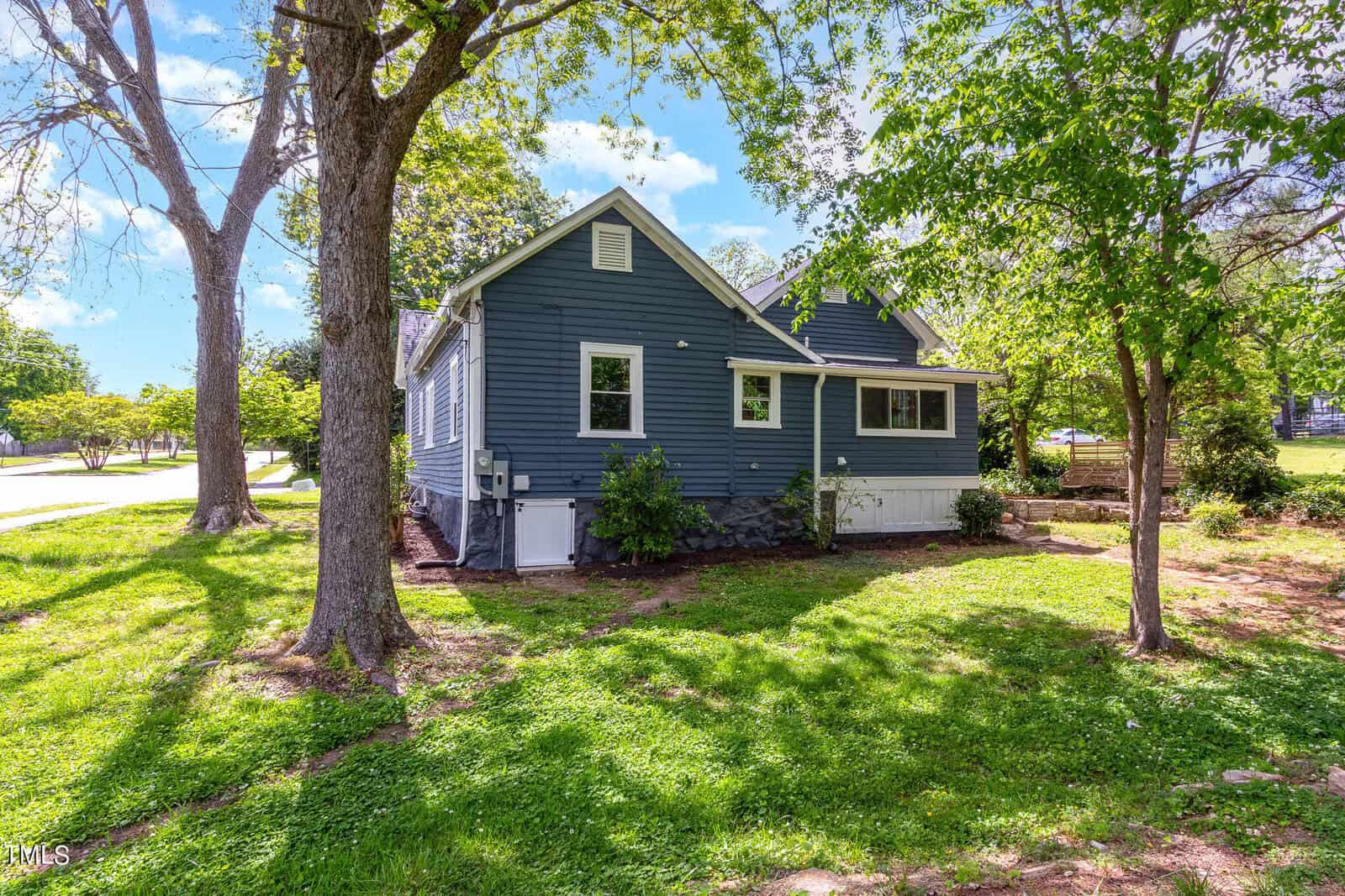 A blue house with white trim set amidst green grass and trees. The sky is bright blue with scattered clouds, and a sidewalk runs beside the lawn.