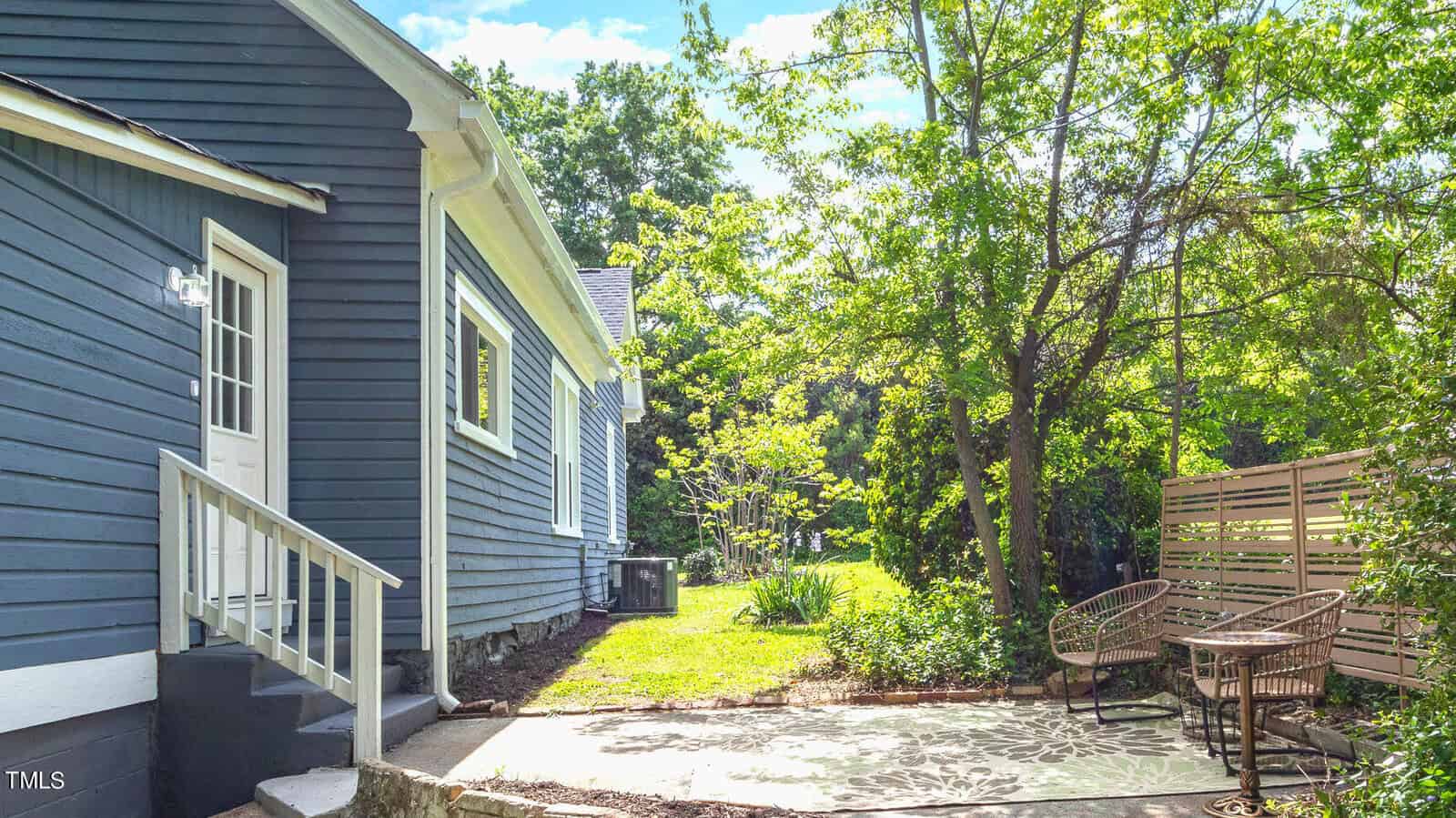 Side view of a blue house with a white trim, featuring a small patio area with two wicker chairs. The area is surrounded by greenery, with tall trees and a wooden fence providing privacy and shade. The sky is clear and sunny.