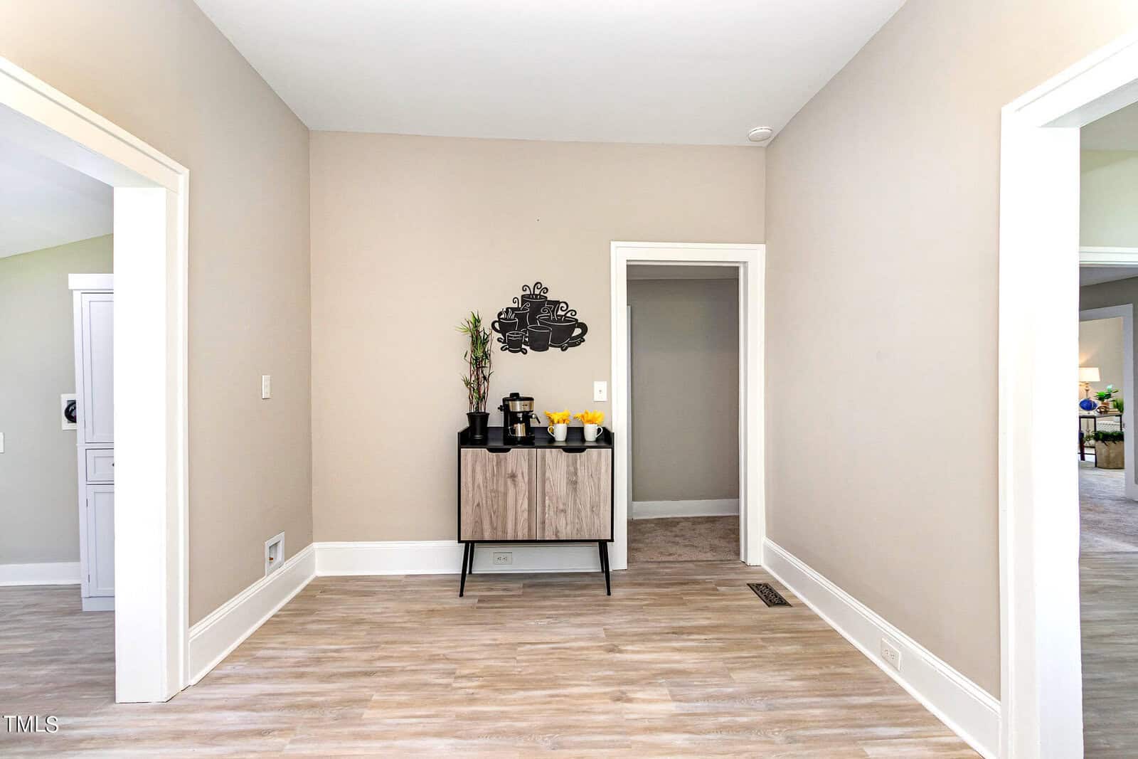 A spacious room with light wood flooring and beige walls. A small cabinet with a decorative sign and potted plant is against one wall. Doors lead to other rooms, and natural light brightens the space.