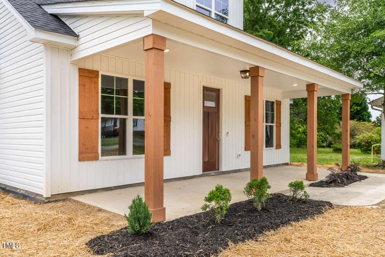 Modern house exterior with white siding and wooden columns. The entrance features a wooden door and windows with shutters. There's a small garden with shrubs lining the front. The roof has dark shingles, and a lawn surrounds the house.