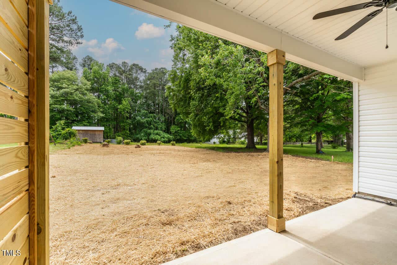 Covered patio with wooden columns opens up to a spacious backyard with a shed. The yard has straw-covered ground and is surrounded by lush green trees under a partly cloudy sky. Ceiling fan visible above the patio.