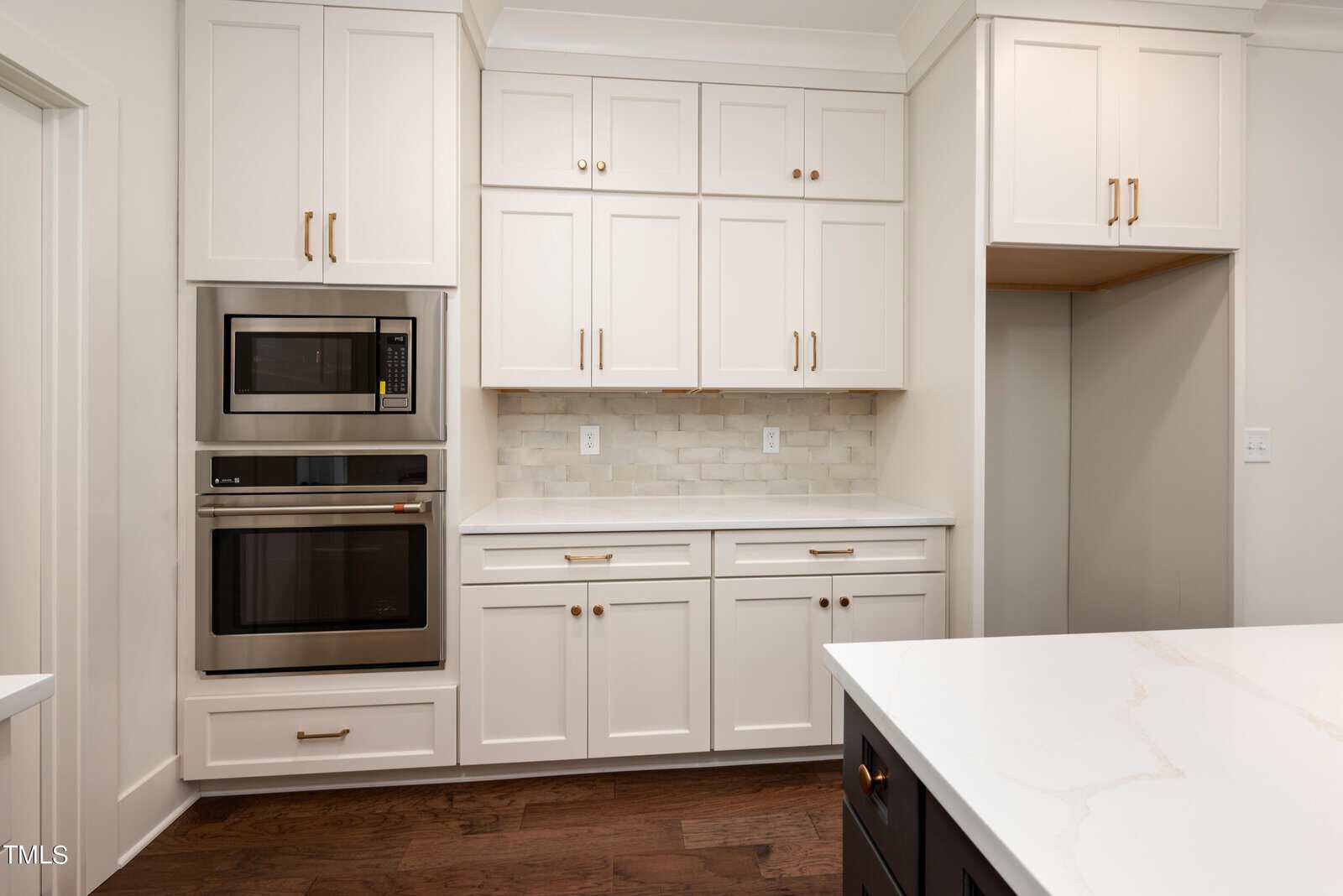 Modern kitchen with white cabinetry, light countertops, and a marble backsplash. Stainless steel double ovens are built into the cabinets. The dark wood floor contrasts with the bright white decor. An island is partially visible in the foreground.