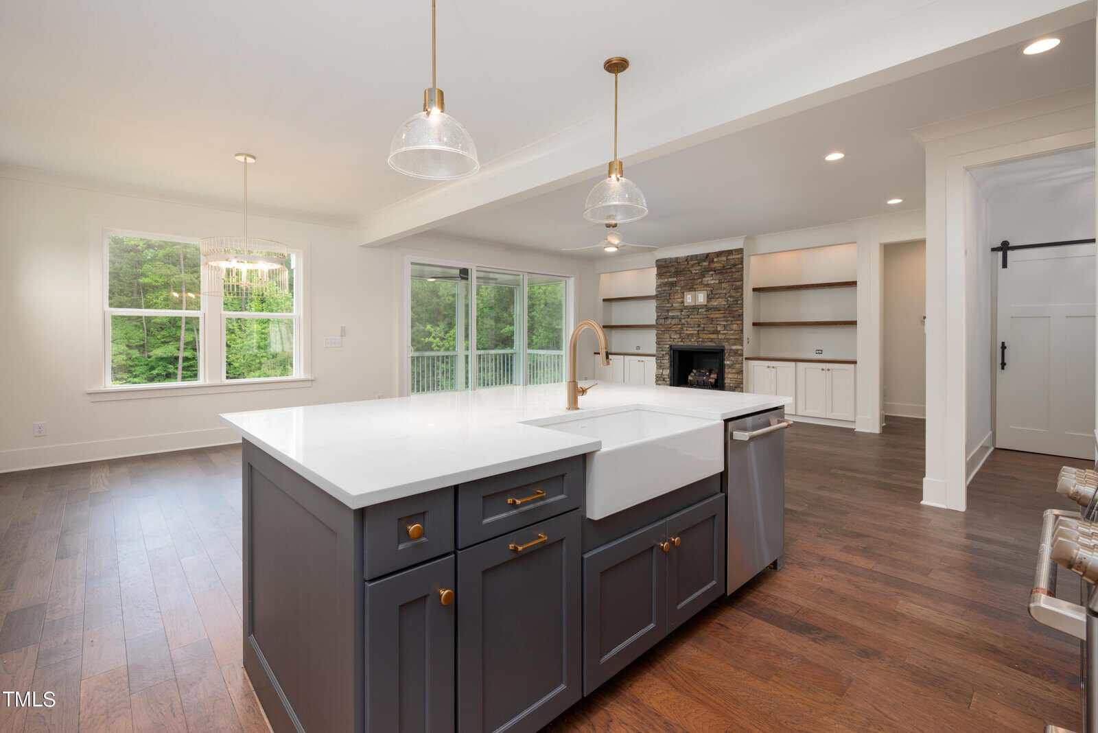 Modern kitchen with a large white countertop island, dark cabinetry, and brass fixtures. Open floor plan leads to a living area with a stone fireplace and built-in shelves. Sliding glass doors overlook a green backyard. Hardwood floors throughout.
